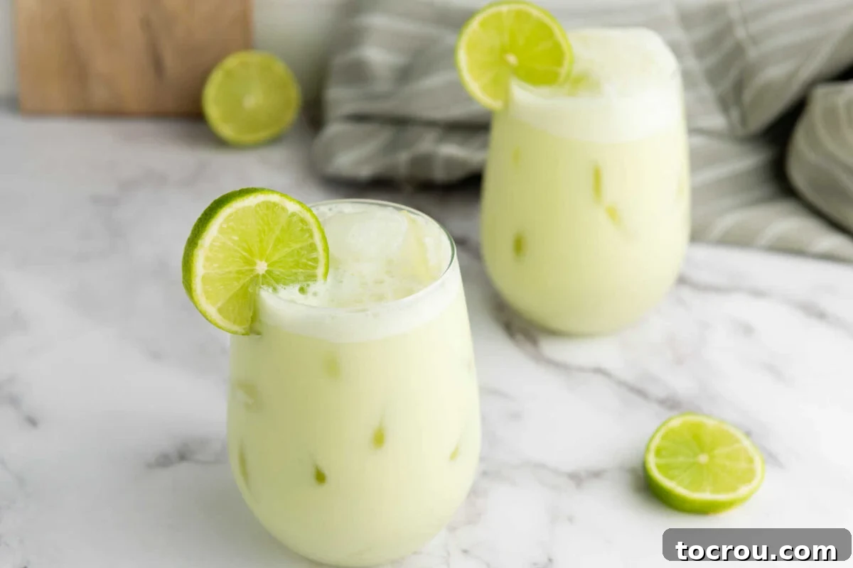 Two tall glasses brimming with creamy, pale green Brazilian limeade, abundant ice cubes, and a fresh lime slice for garnish, set against a bright background.