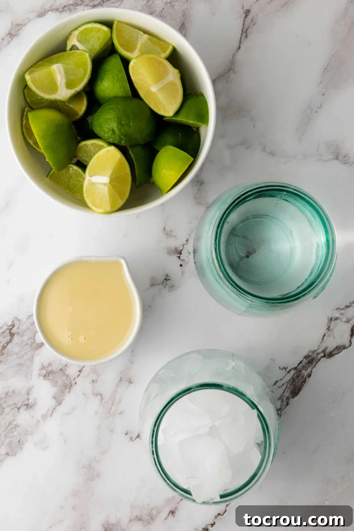 A collection of essential ingredients laid out for Brazilian limeade preparation: fresh green limes, a can of sweetened condensed milk, a pitcher of water, and a bowl of ice cubes.