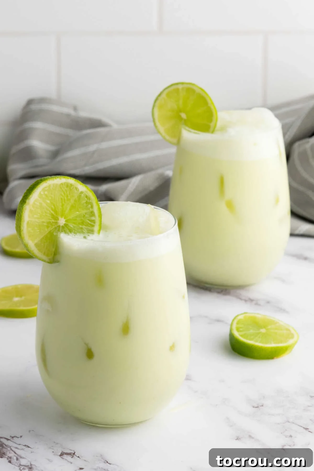 Two clear glasses filled with creamy, light green Brazilian limeade and ice, garnished with vibrant green lime slices, ready to be enjoyed.