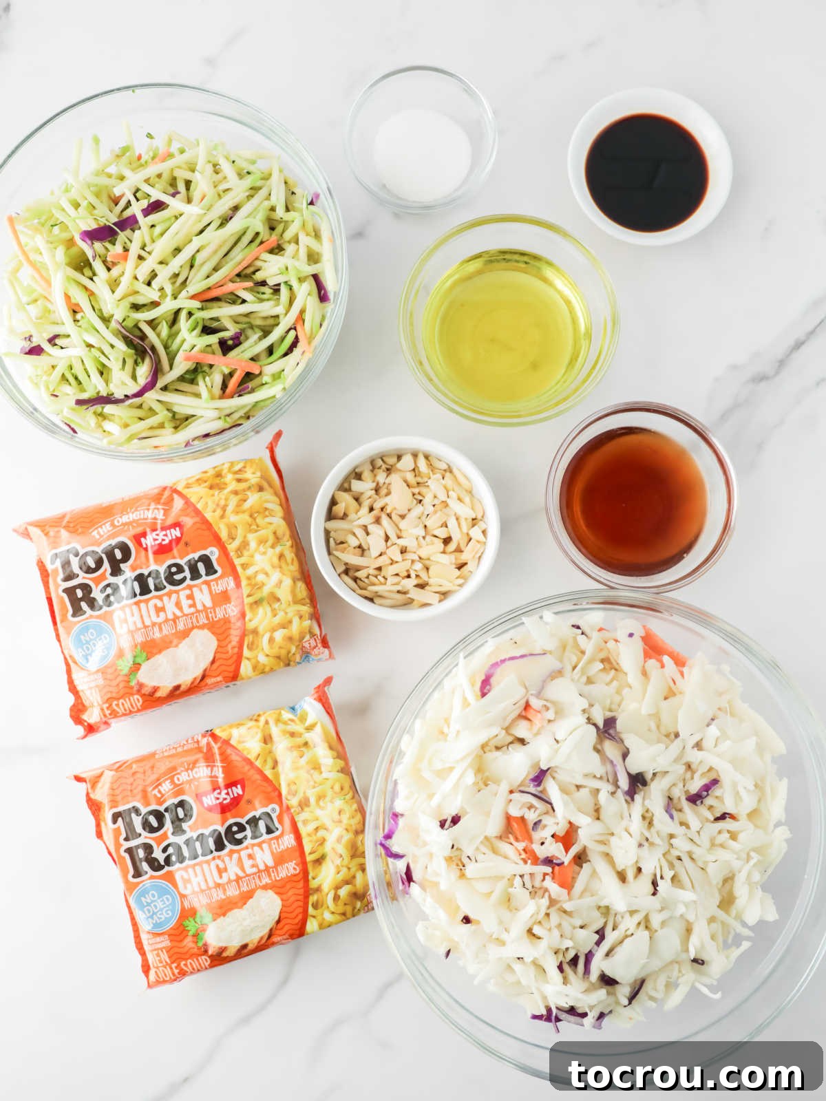 Crunchy Ramen Slaw 3 A flat lay photograph showcasing all the fresh ingredients needed for ramen noodle coleslaw: a large bag of classic coleslaw mix, a bag of shredded broccoli slaw mix, two packages of instant ramen noodles, a small bowl of slivered almonds, and individual glass bowls containing soy sauce, olive oil, granulated sugar, and rice vinegar, all neatly arranged on a rustic wooden surface.