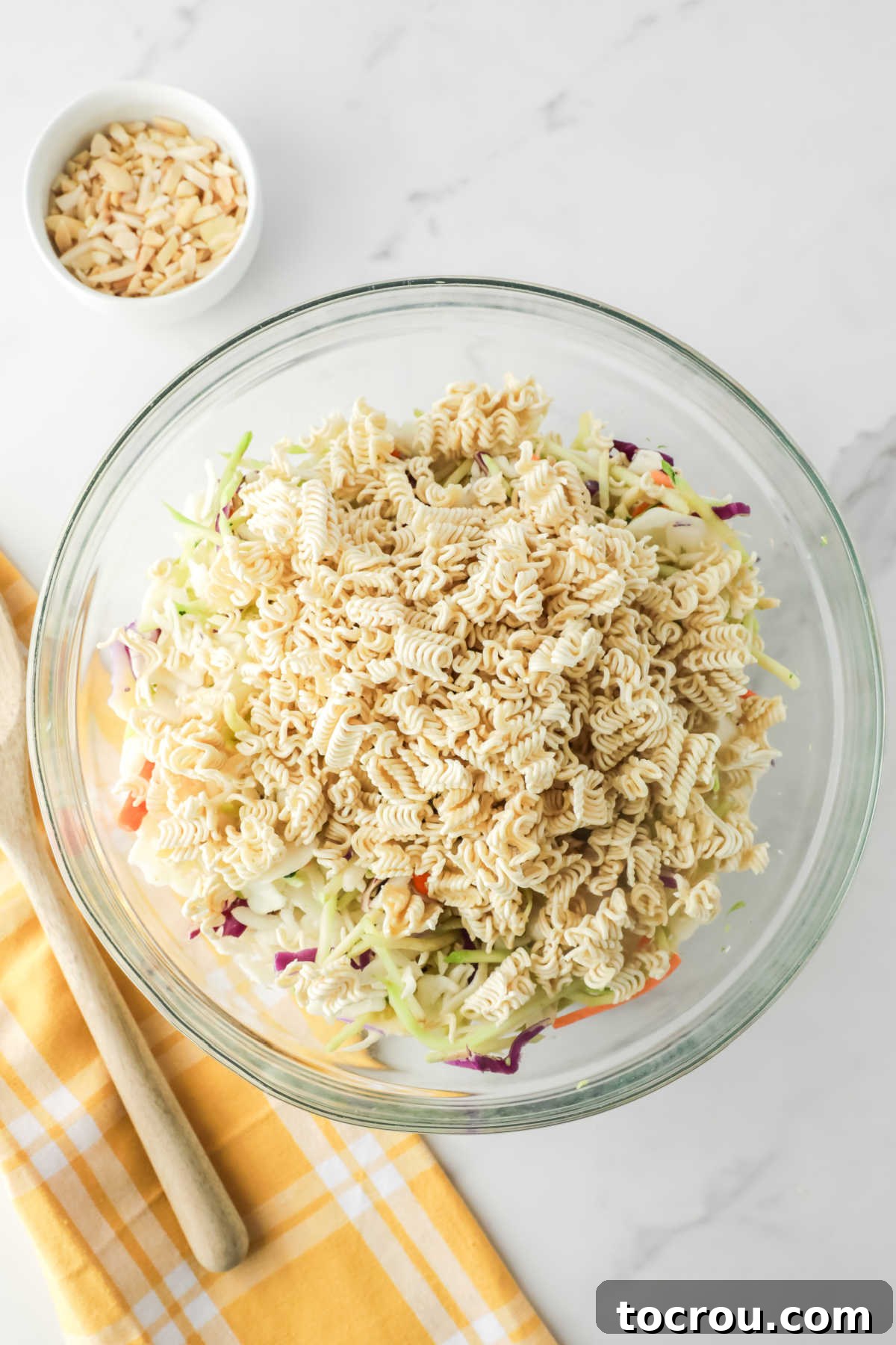Crunchy Ramen Slaw 4 A large mixing bowl filled with the combined shredded vegetables from classic coleslaw and vibrant broccoli slaw mixes, with visibly broken pieces of uncooked ramen noodles scattered generously throughout the colorful blend, ready to be coated in the flavorful dressing.