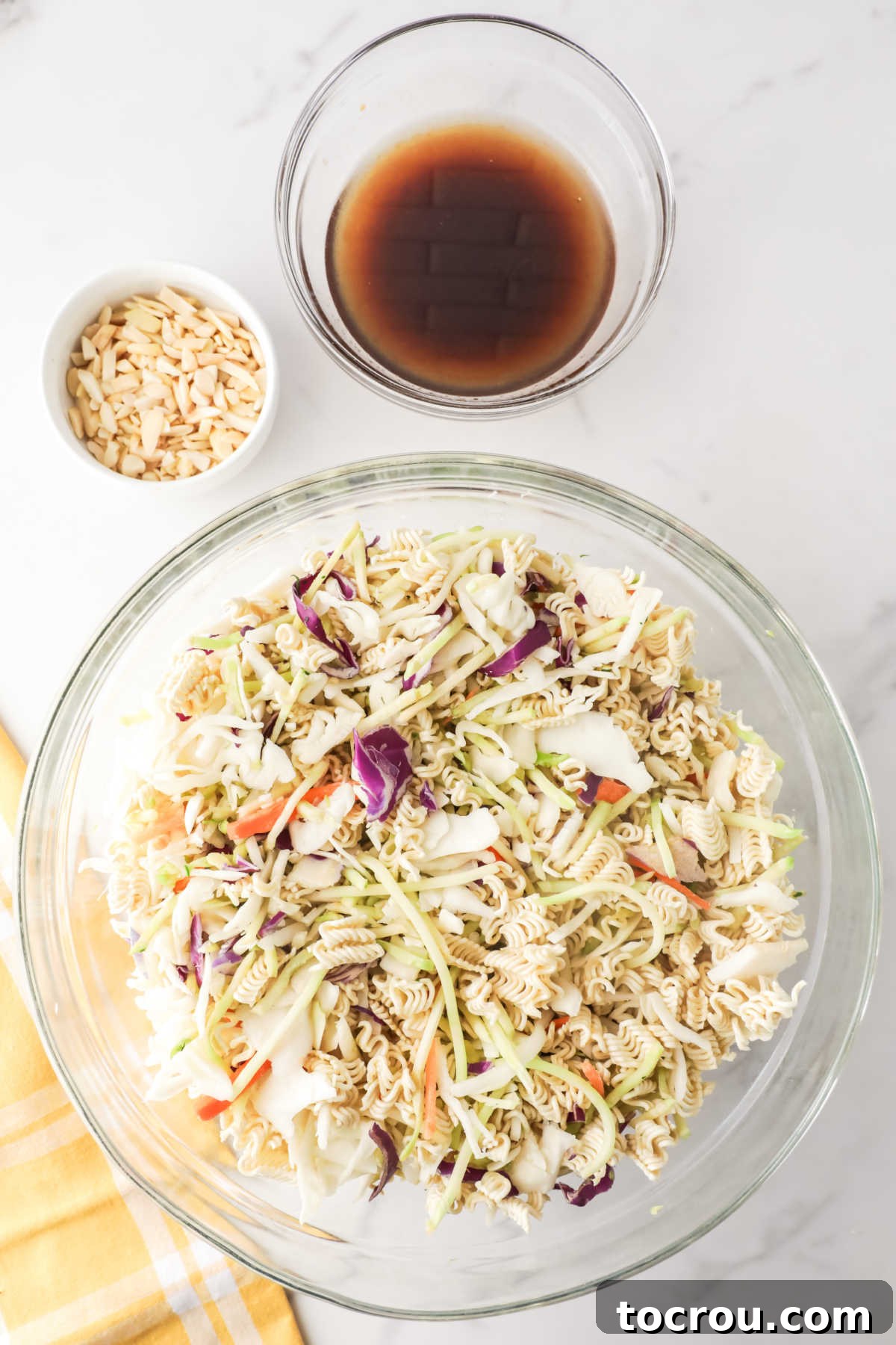 Crunchy Ramen Slaw 5 A small glass bowl containing the freshly prepared homemade soy vinaigrette dressing is strategically positioned next to a large mixing bowl. The mixing bowl holds the combined shredded coleslaw and broccoli slaw, along with broken pieces of ramen noodles, all meticulously arranged and ready to be mixed together to create the vibrant ramen noodle coleslaw.