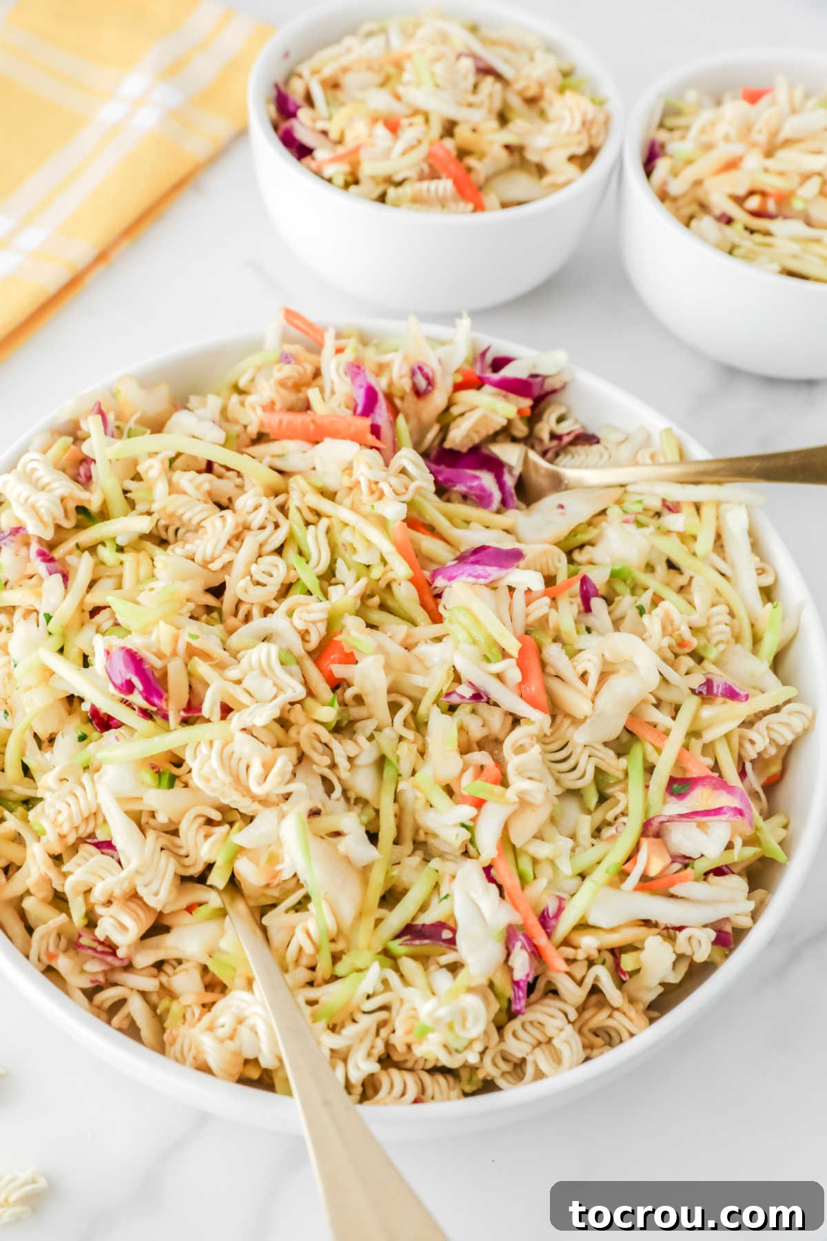 Crunchy Ramen Slaw 6 A large, aesthetically pleasing serving bowl filled to the brim with freshly prepared ramen noodle coleslaw, showcasing the colorful medley of shredded green cabbage, vibrant broccoli, bright carrots, an abundance of crunchy ramen noodles, and delicate slivered almonds. Two smaller, individual serving bowls of the same delicious slaw are artfully arranged in the soft-focused background, indicating a meal ready to be enjoyed.