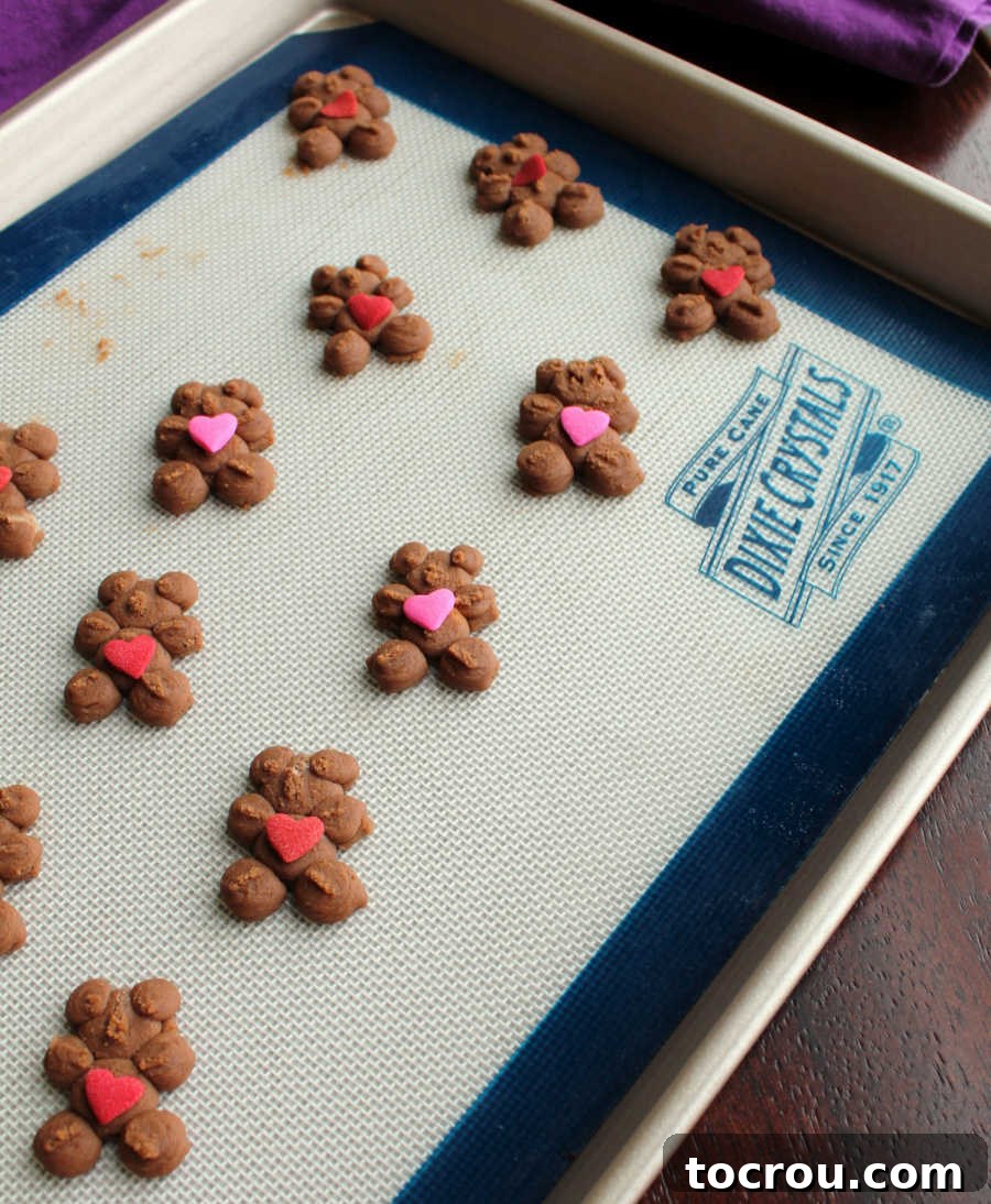 A baking sheet filled with unbaked bear-shaped chocolate spritz cookies, each topped with vibrant red and pink heart candies, ready for the oven.