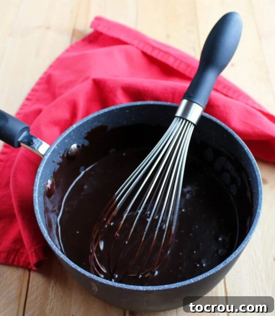 A saucepan on the stove, holding a warm, rich ganache-style frosting, with a whisk resting inside, showing its smooth consistency.