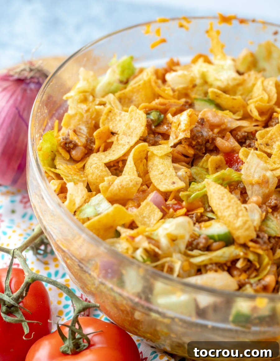 A family-sized Frito Taco Salad, packed with fresh ingredients and homemade dressing. A large glass serving bowl brimming with colorful taco salad, featuring crunchy Fritos, fresh cucumbers, hearty beans, ripe tomatoes, crisp lettuce, and a generous drizzle of homemade Catalina dressing.