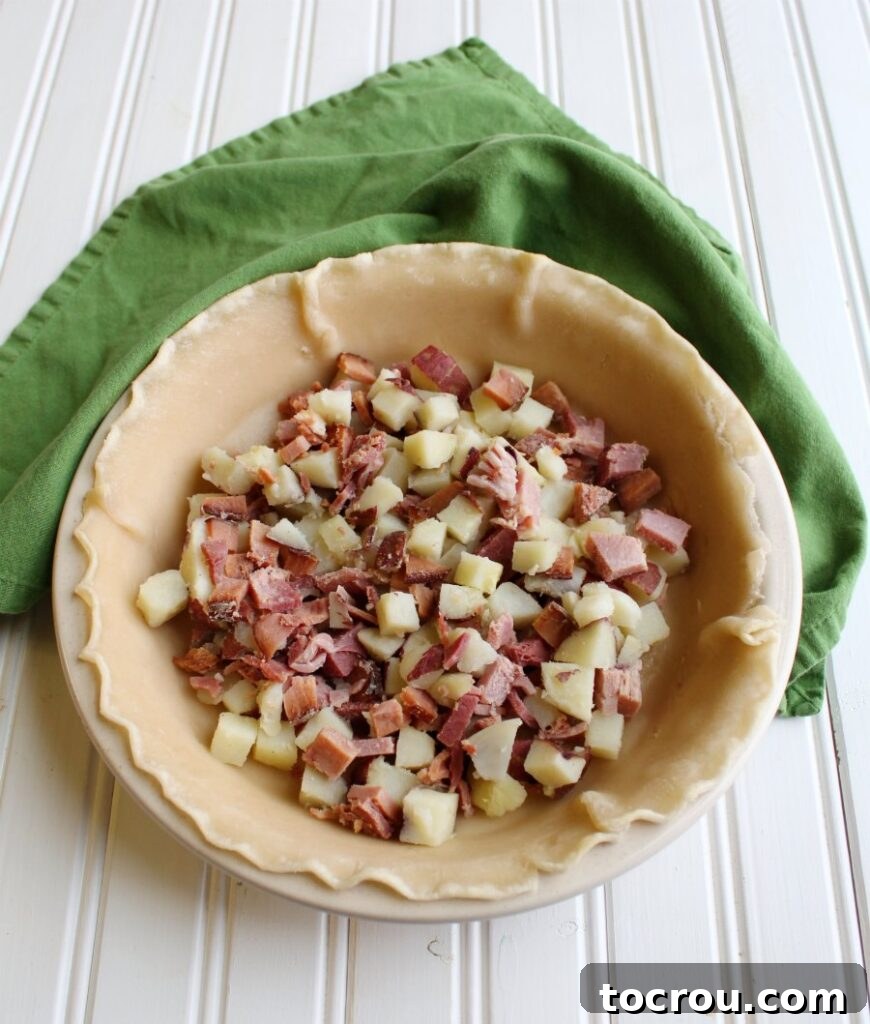 An unbaked pie shell filled generously with diced ham and tender potatoes, ready for the egg custard.
