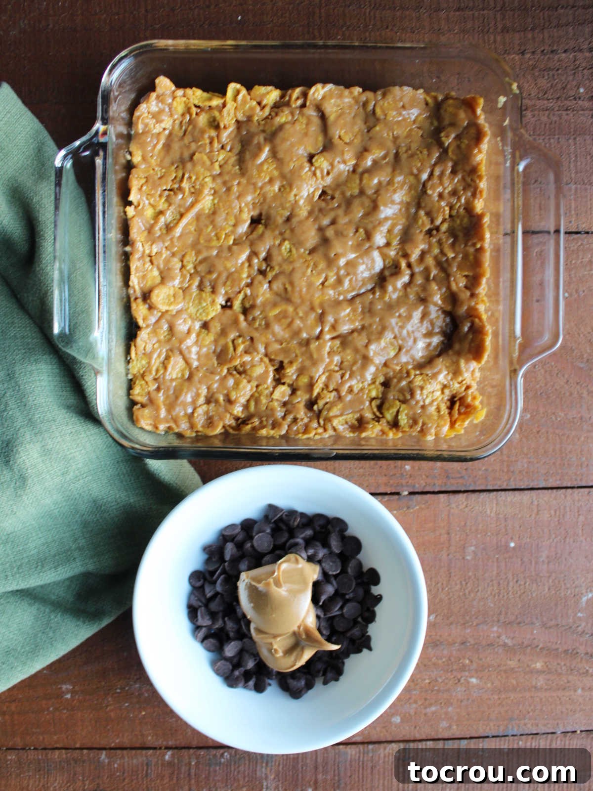 Ready for the chocolate topping. Peanut butter cornflake mixture pressed into a square baking pan, with chocolate chips and peanut butter nearby, ready for the topping.