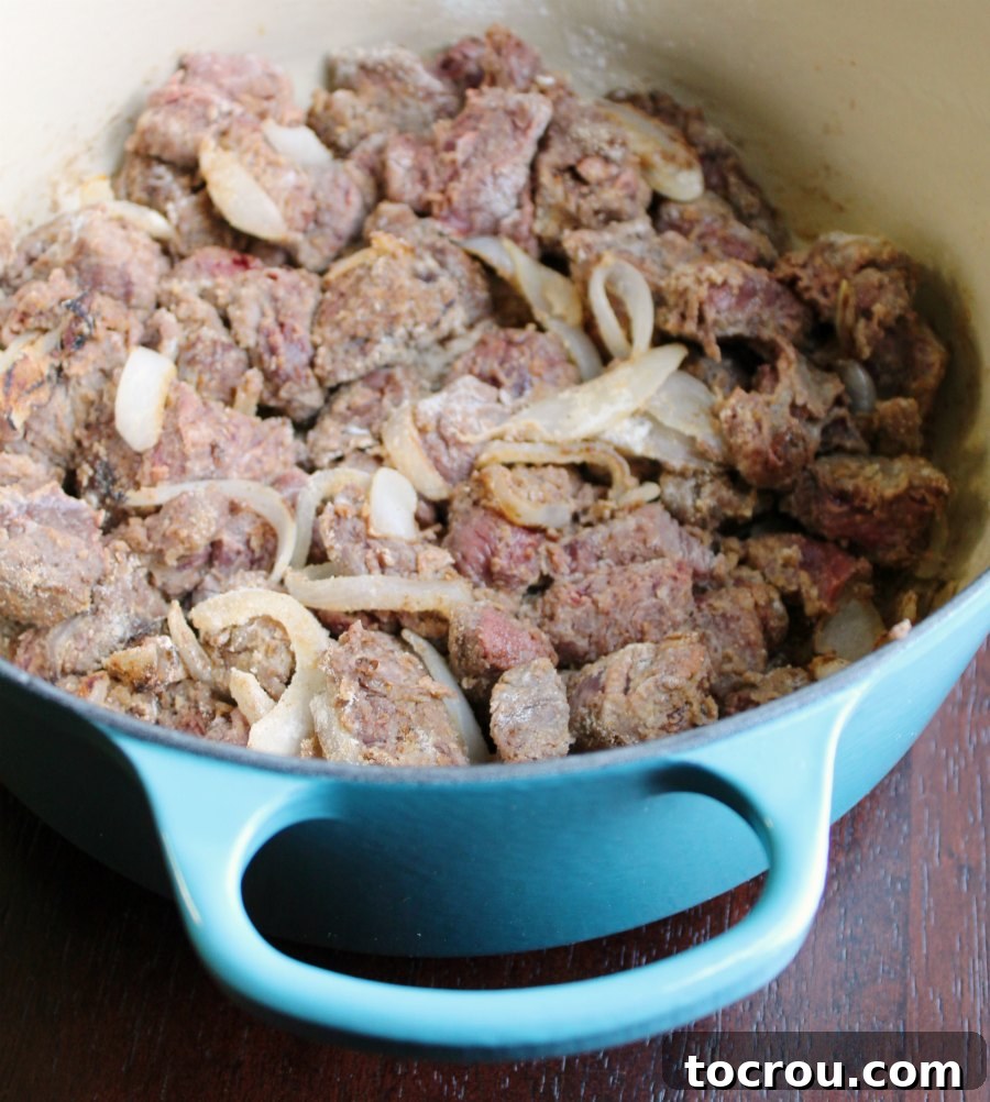 Close-up of beef stew meat and sliced onions being sautéed in a vibrant enameled Dutch oven.