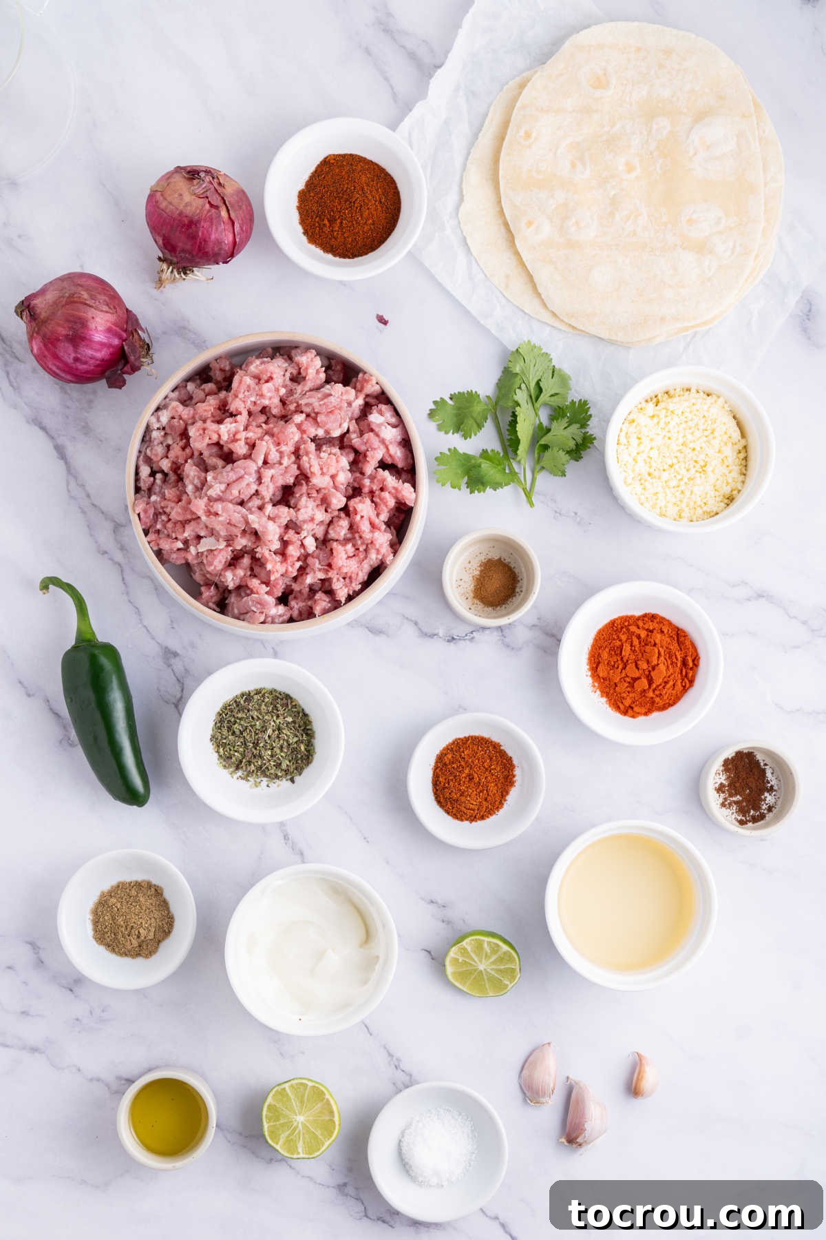 A selection of raw ingredients laid out on a wooden table, including ground pork, various spices in small bowls, sliced jalapenos, chopped onions, and a stack of corn tortillas, all ready for preparing fresh chorizo and tacos.