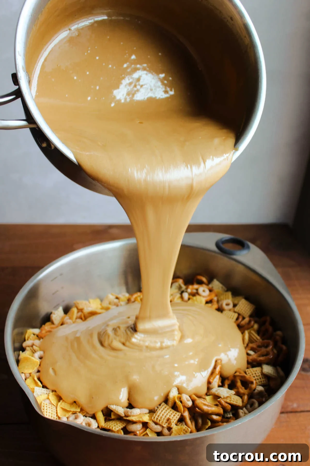 Pouring hot caramel mixture over a bowl of cereal, pretzels and peanuts.
