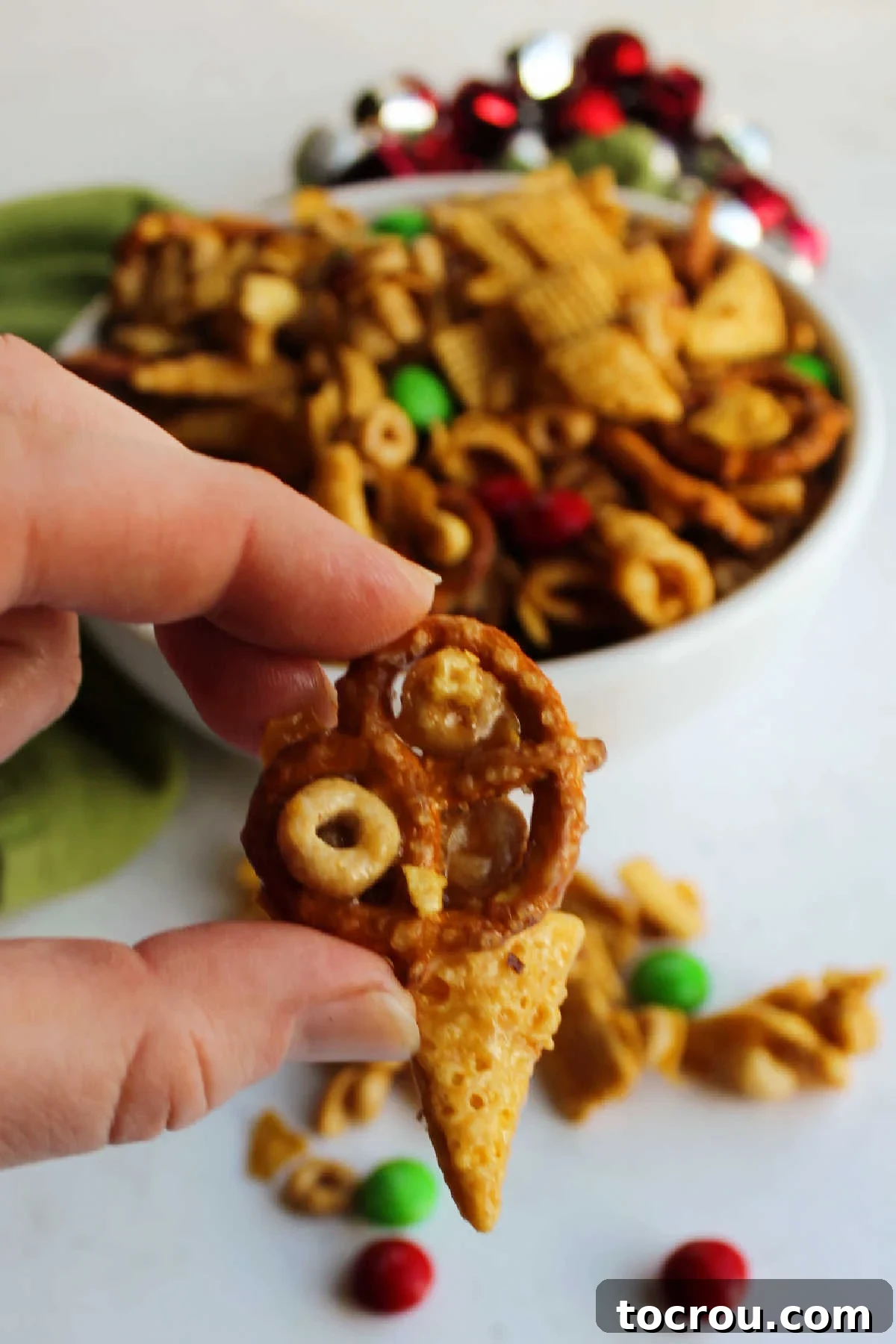 Hand holding a cluster of cheerios, a pretzel and a bugle all held together with a crunchy caramel mixture.