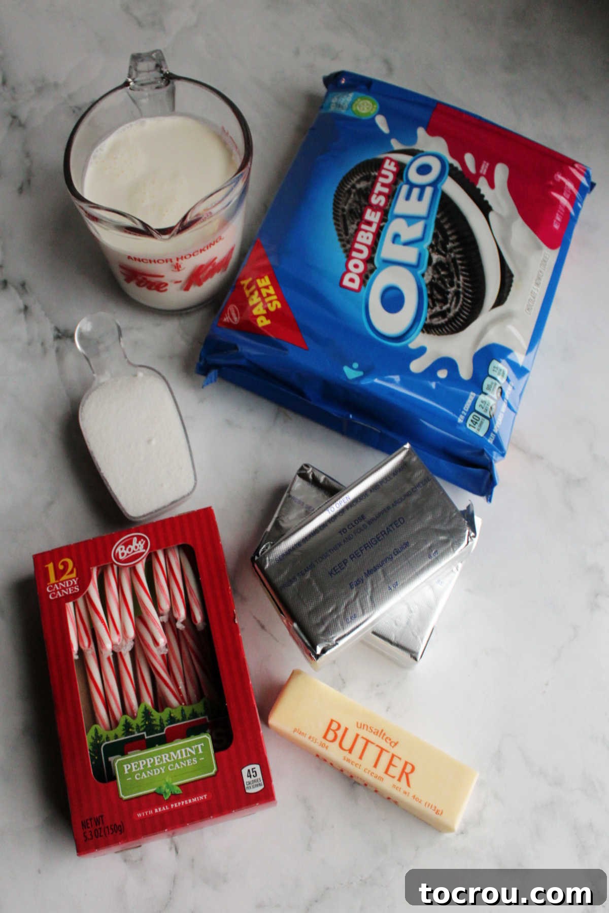 Key ingredients for no bake cheesecake including Oreos, cream cheese, heavy cream, butter, sugar and candy canes laid out on a white surface.