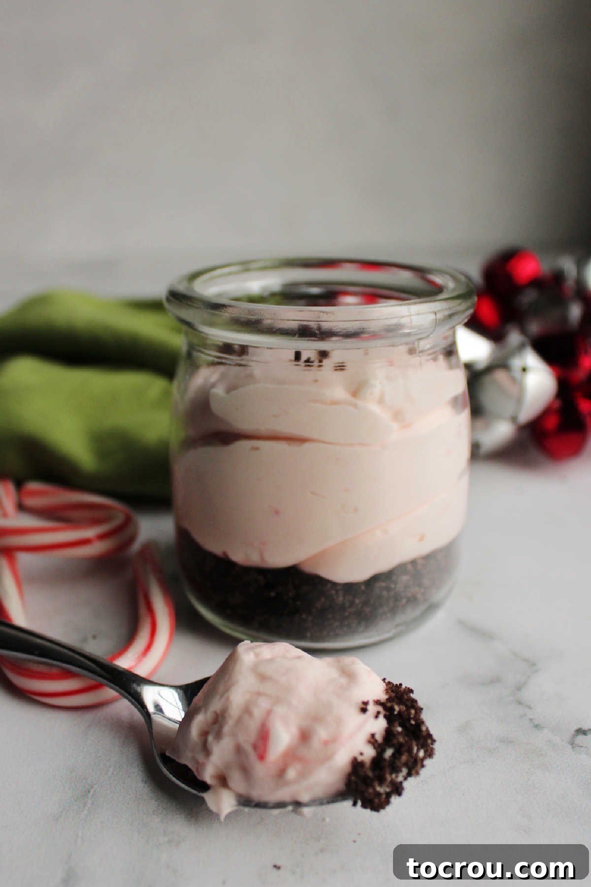 A spoon holds a delightful bite of creamy peppermint cheesecake filling and Oreo crumbs, presented in front of a jar filled with the dessert, highlighting the texture and layers.