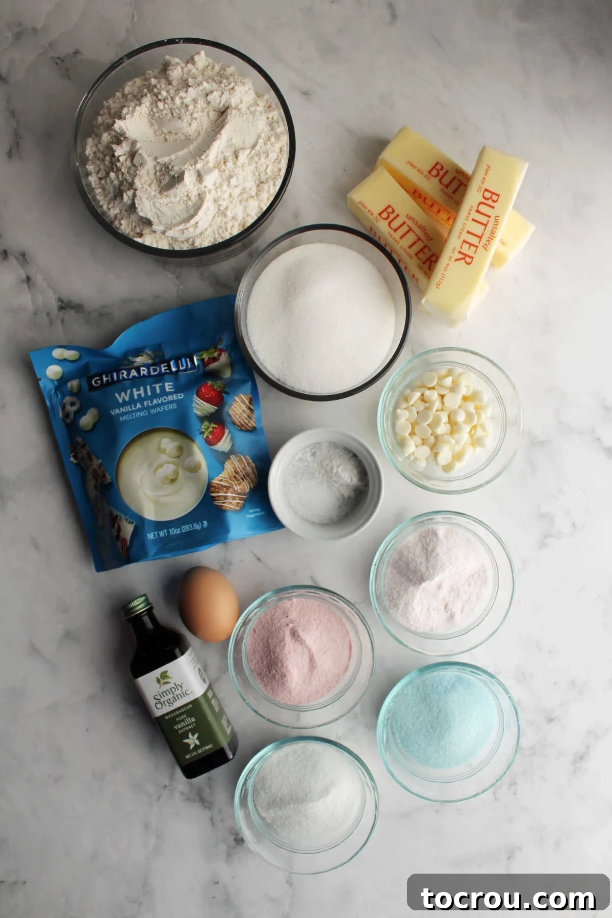 An array of ingredients neatly arranged, including flour, softened butter, granulated sugar, baking powder, salt, a fresh egg, vanilla extract, various Jell-O powder boxes, white chocolate melting disks, and white chocolate baking chips, all prepared for making delicious paw print cookies.