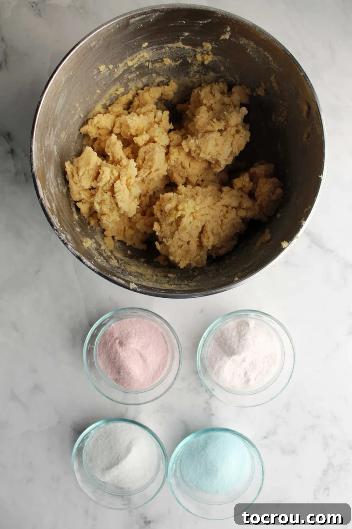 A mixer bowl containing prepared sugar cookie dough, positioned next to four small bowls each filled with different colors of Jell-O powder, ready for mixing.