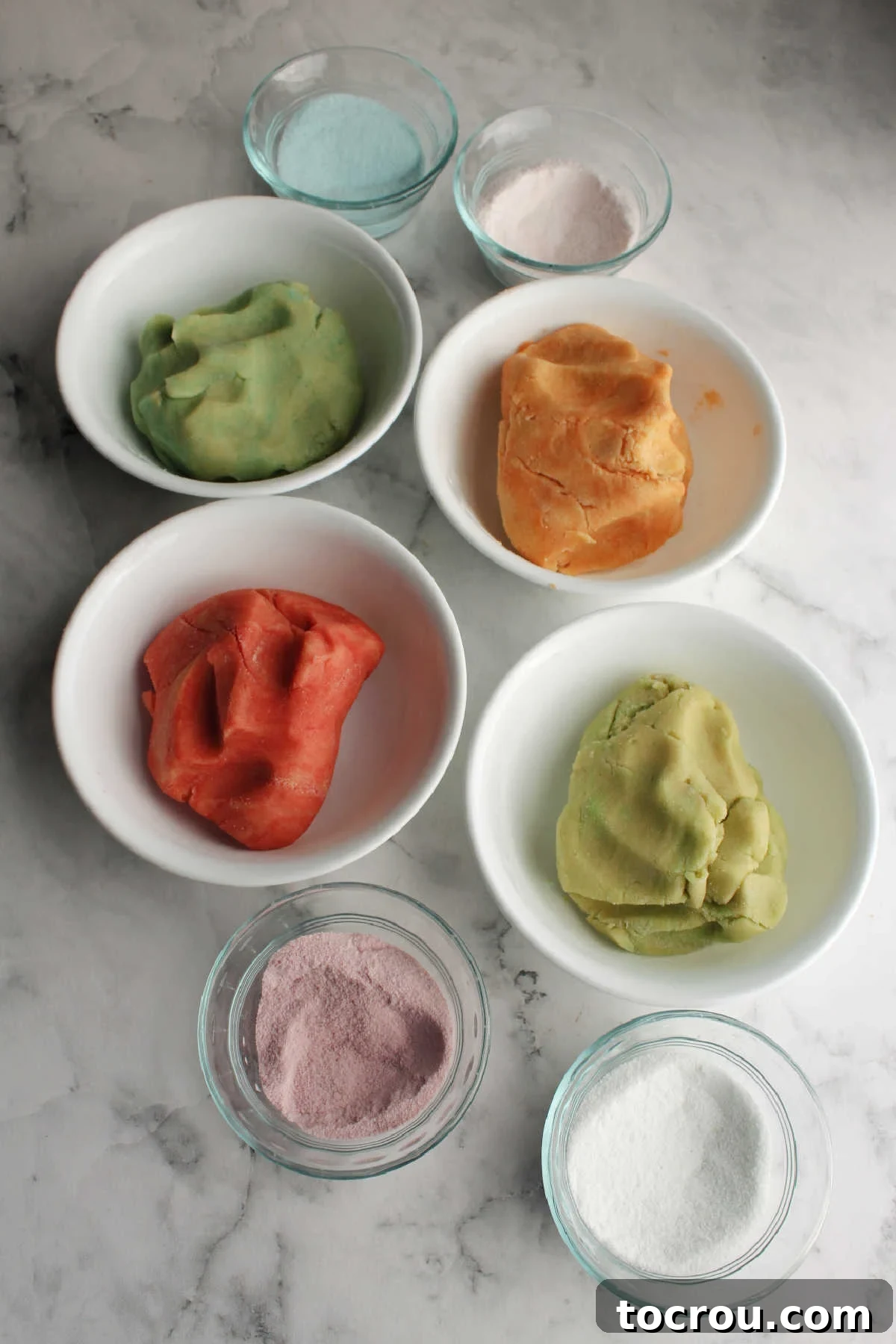 Four separate small bowls, each containing a different vibrant color of cookie dough, with smaller companion bowls holding extra Jell-O powder for rolling.