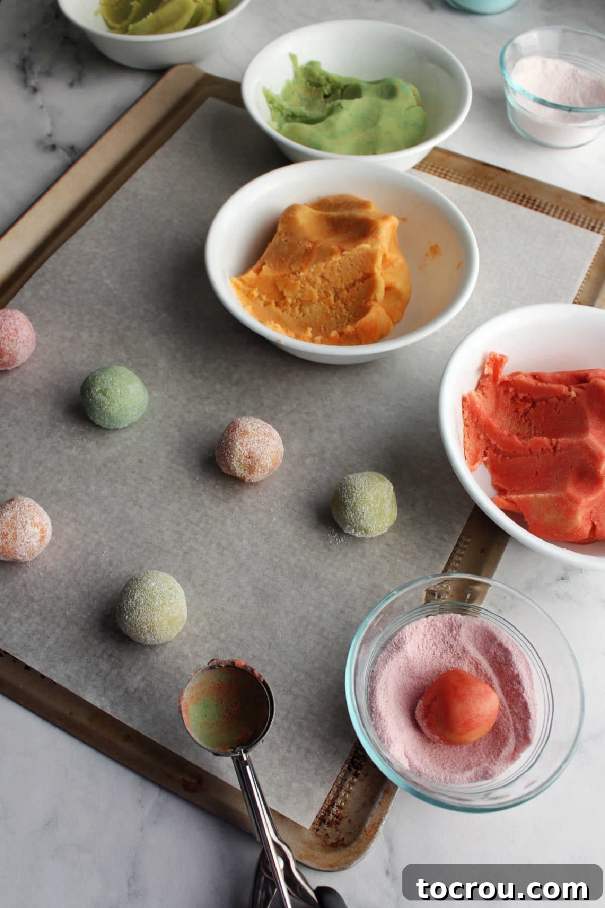 A baking enthusiast skillfully uses a small cookie scoop to portion out dough, then rolls it in additional Jell-O powder to enhance its color and flavor before baking.