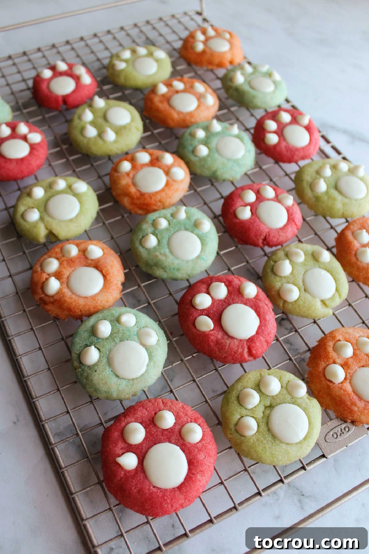 A vibrant collection of colorful cookies, each adorned with white chocolate paw decorations, cooling on a wire rack, showcasing their irresistible charm.