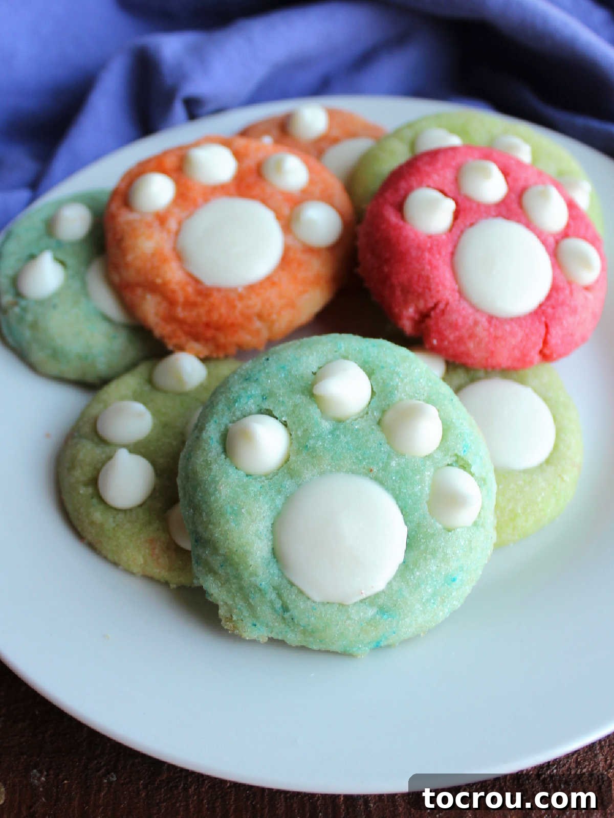 A beautifully arranged plate featuring an assortment of blue, pink, orange, and green fruity cookies, each decorated with pristine white chocolate paw prints, perfectly ready for serving and enjoyment.