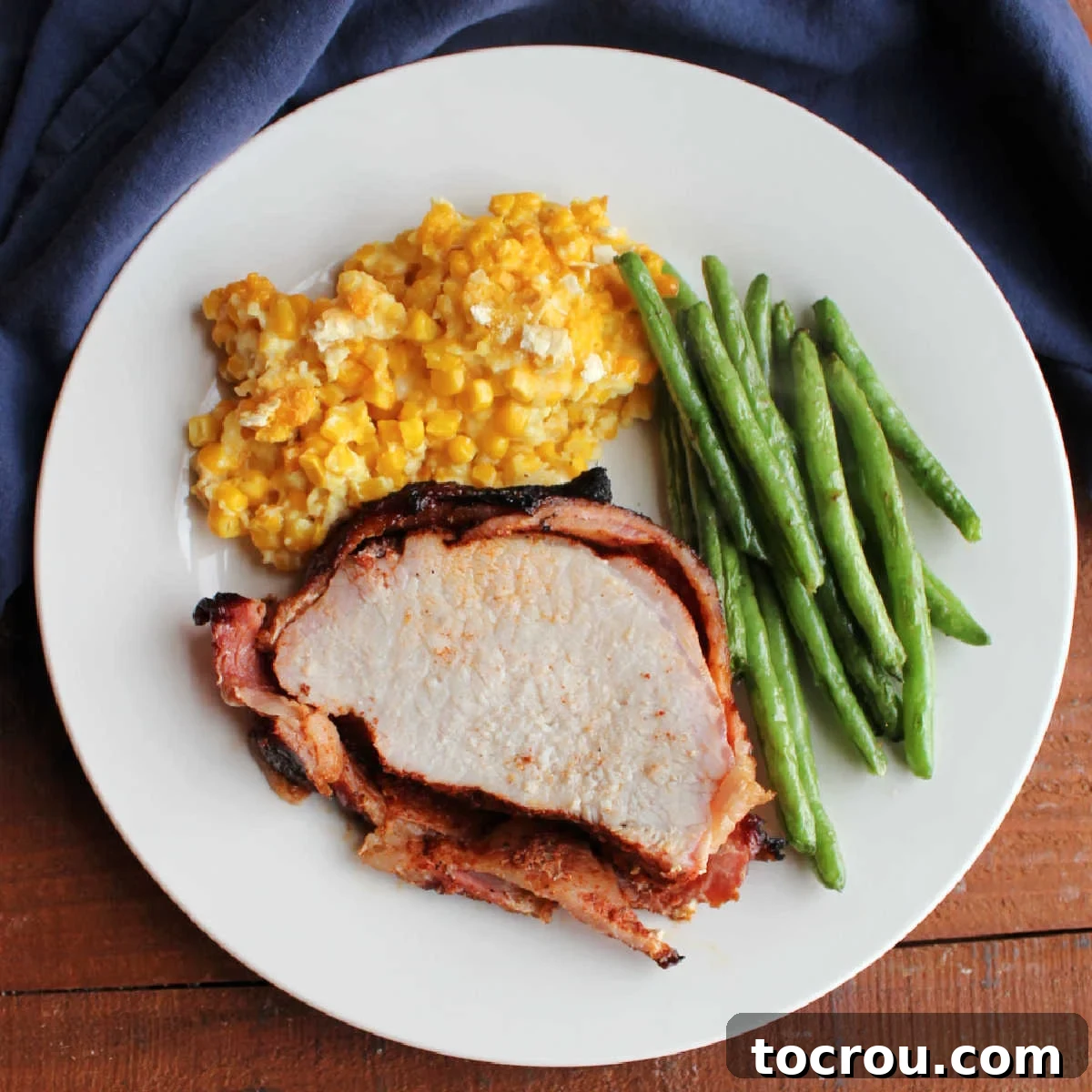Dinner plate with bacon wrapped smoked pork loin, roasted green beans, and scalloped corn on it, ready to eat.