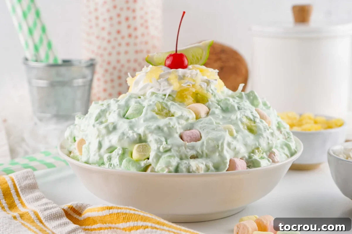 Serving bowl of seafoam salad with pale green color and fluffy texture dotted with colorful marshmallows and topped with whipped cream and a cherry.