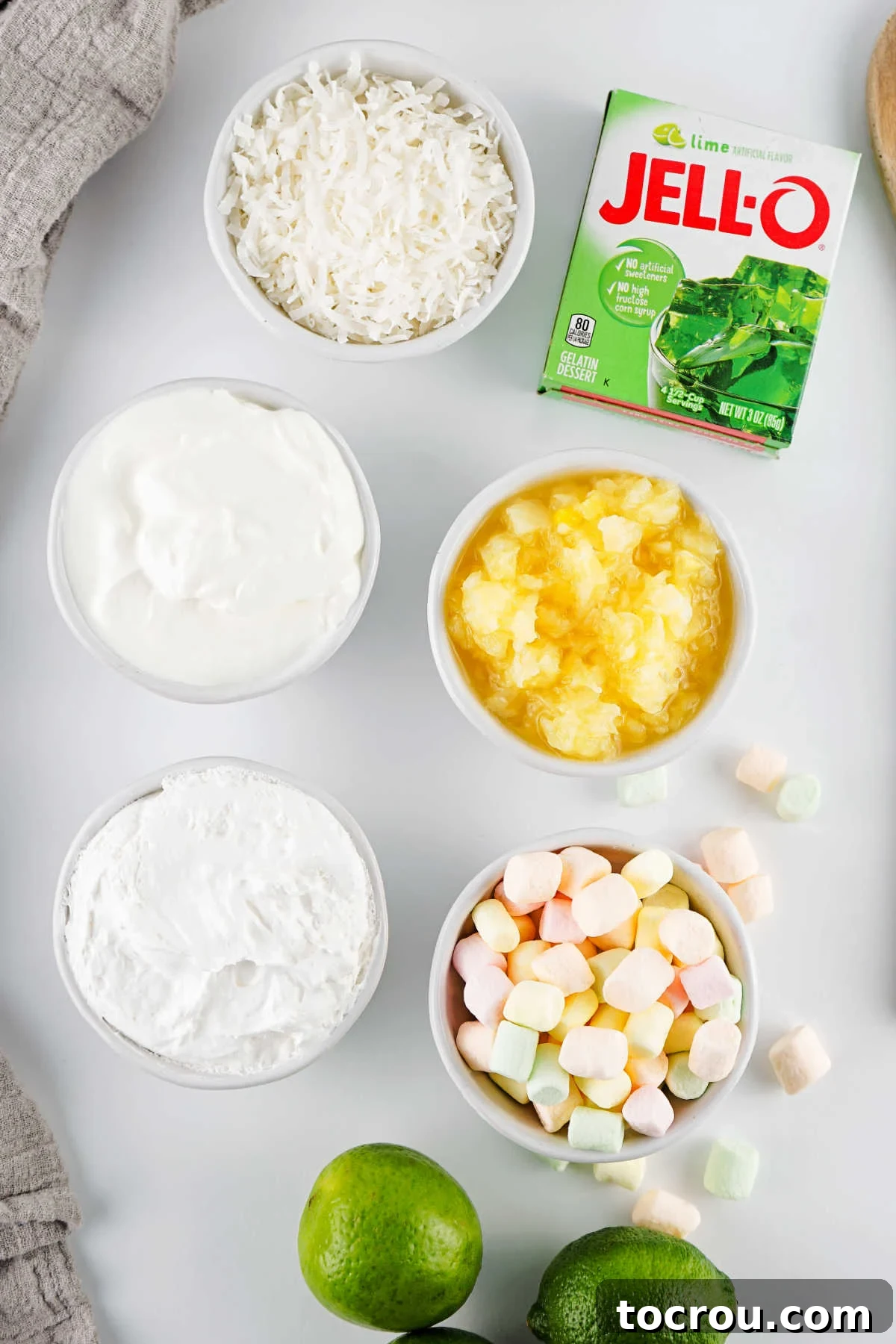 Ingredients including lime jello, crushed pineapple, pastel marshmallows, sour cream, cool whip, and shredded coconut ready to be made into lime fluffy seafoam salad. 