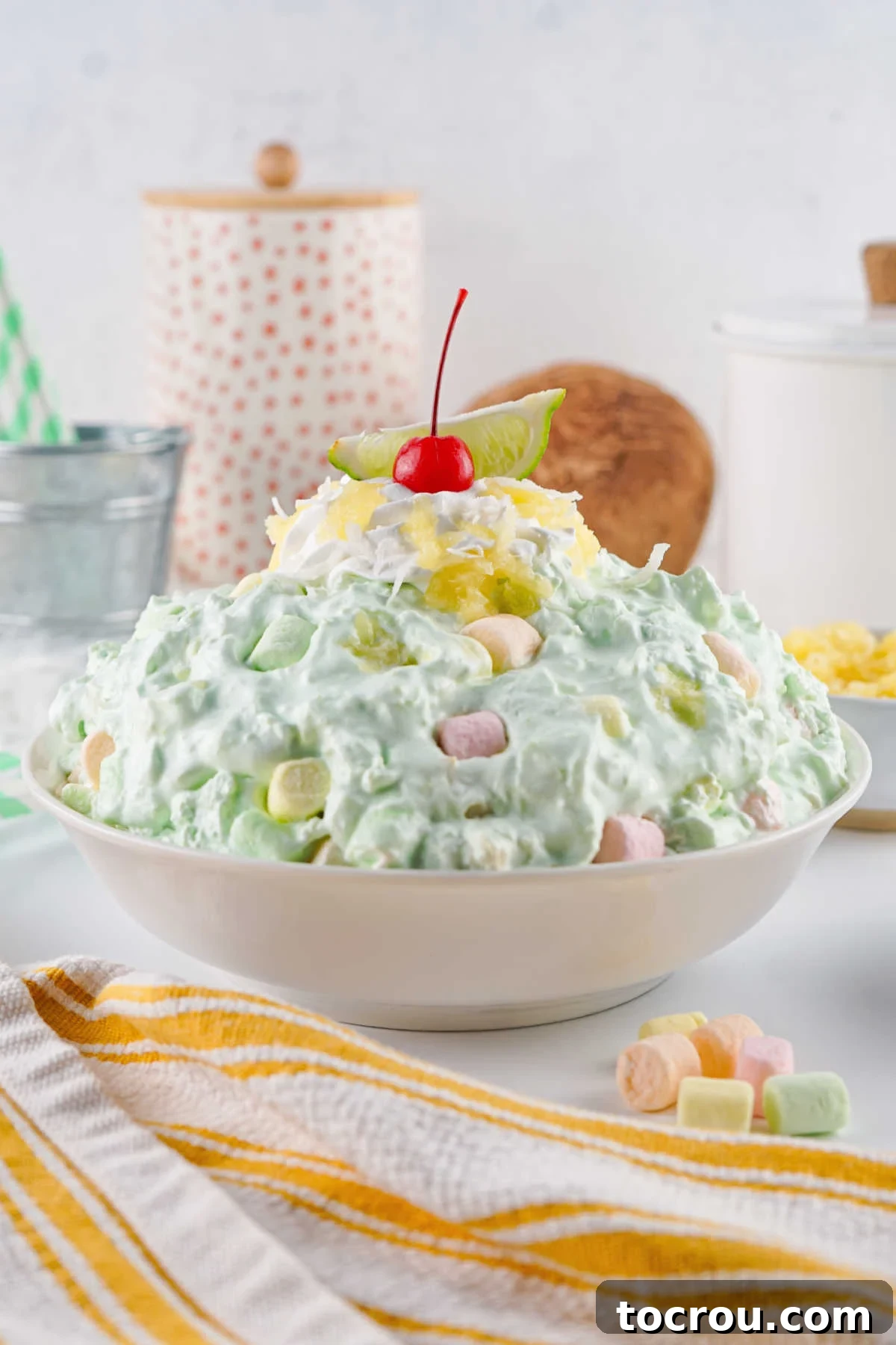 Large bowl of pale green seafoam salad with lime wedge, whipped cream, marshmallows, and cherry on top.