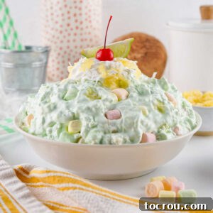Serving bowl filled with lime jello fluff salad with pale green color, pastel marshmallows, whipped cream and a cherry on top.