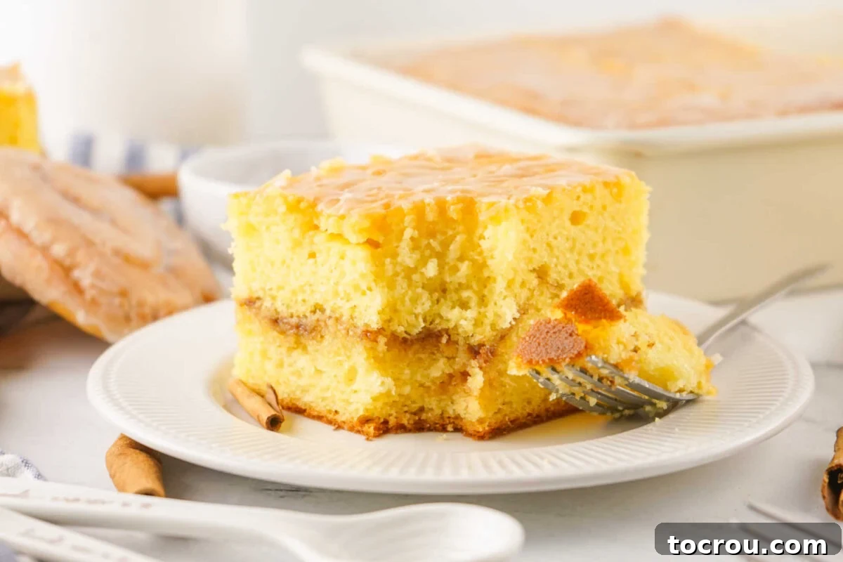 Fork getting a bite of honey bun cake with a distinct layer of cinnamon and brown sugar in the middle of a rich yellow cake, topped with a thin white glaze.