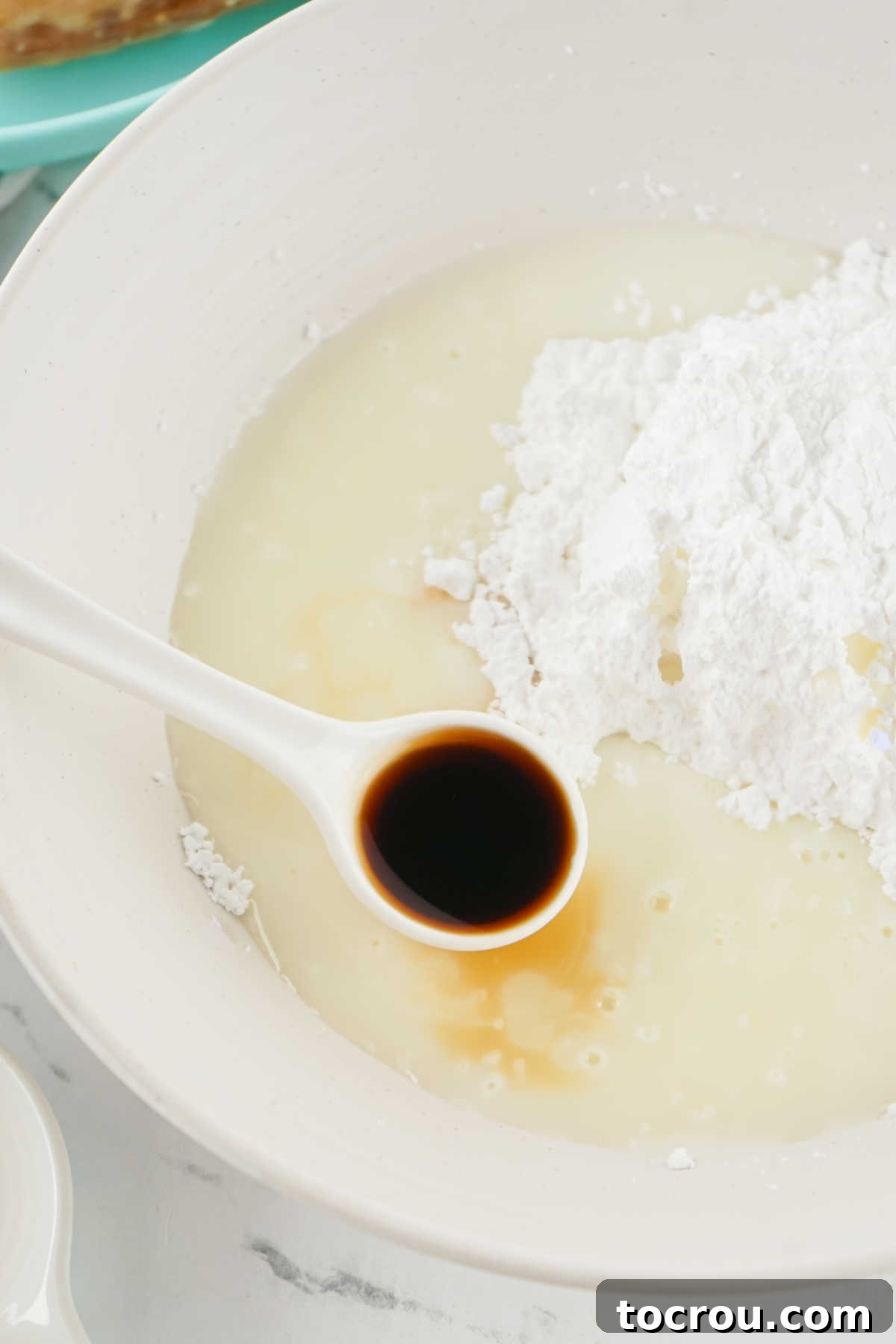 Adding vanilla extract to a bowl containing milk and powdered sugar, mixing to create a smooth glaze for the cake.