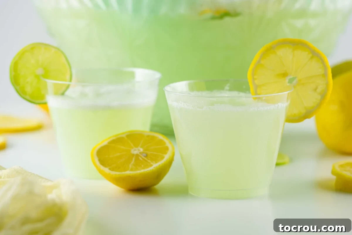 Two glasses of pale green lemon lime sherbet punch garnished with a lemon and lime slice in front of punch bowl with remaining punch.