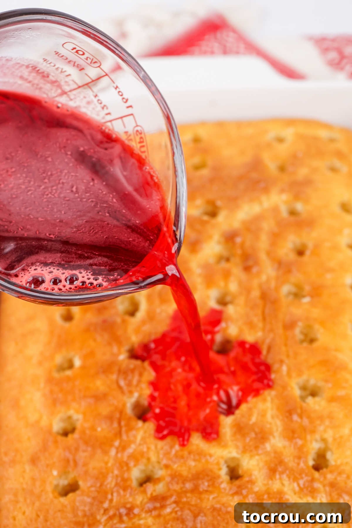 Ruby Cranberry Infusion Cake 5 Pouring warm cranberry jello mixture over the top of cake with holes poked in it.