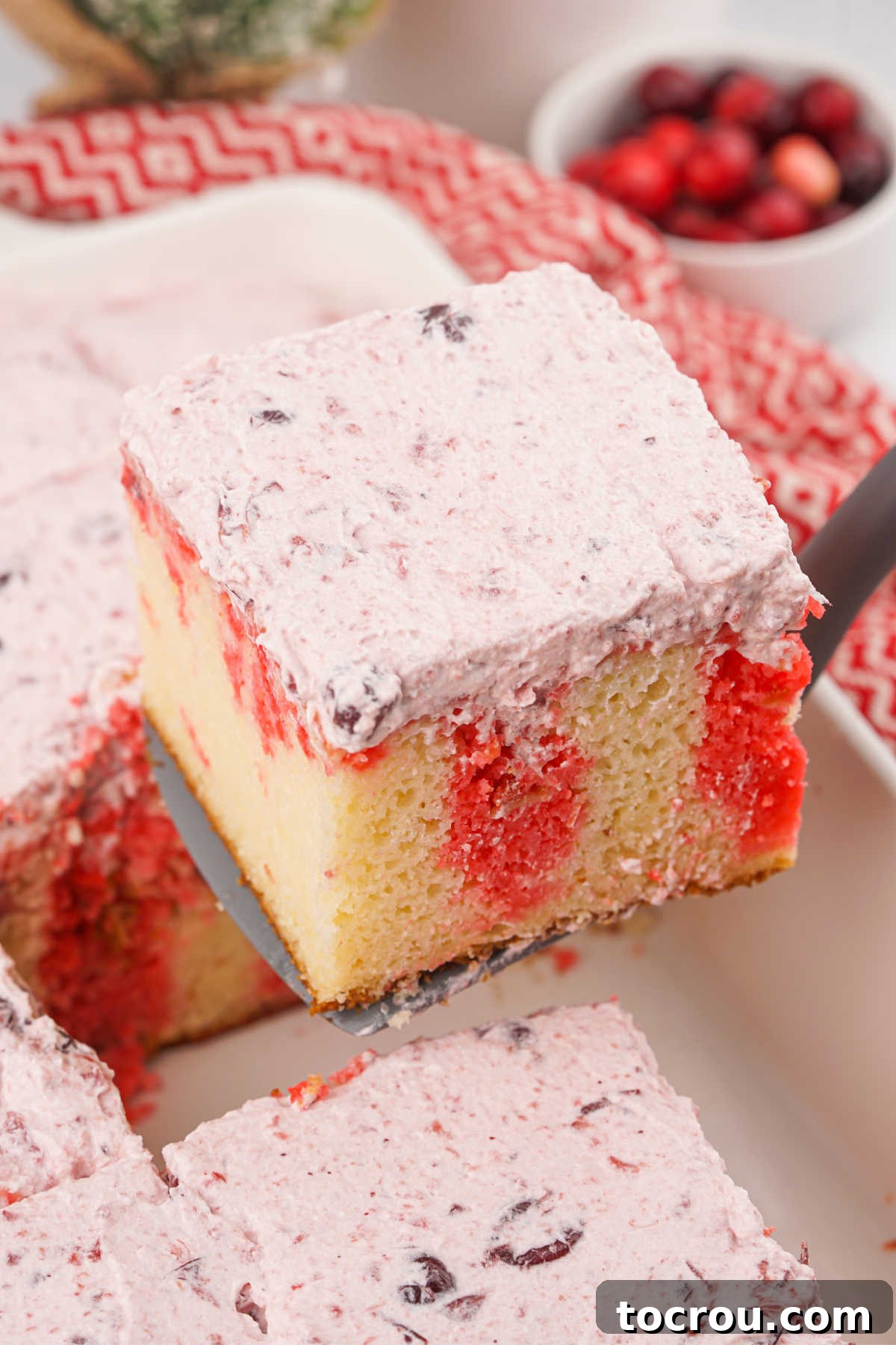 Ruby Cranberry Infusion Cake 8 Spatula lifting piece of jello poke cake out of pan showing vanilla cake, stripes of cranberry jello, and cranberry frosting on top.