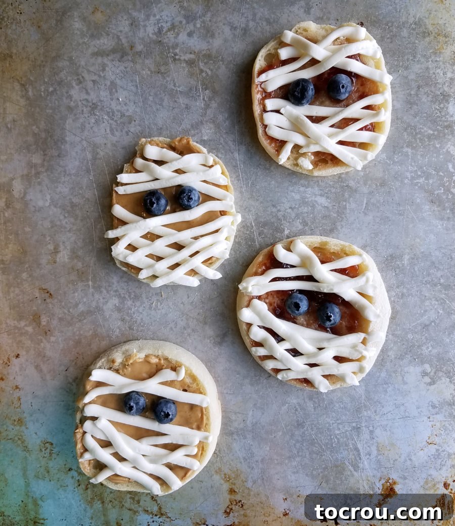Delicious and Spooky English Muffin Mummies Ready to Eat Finished English muffin mummies with peanut butter, jelly, cream cheese, and blueberry eyes, arranged on a plate.