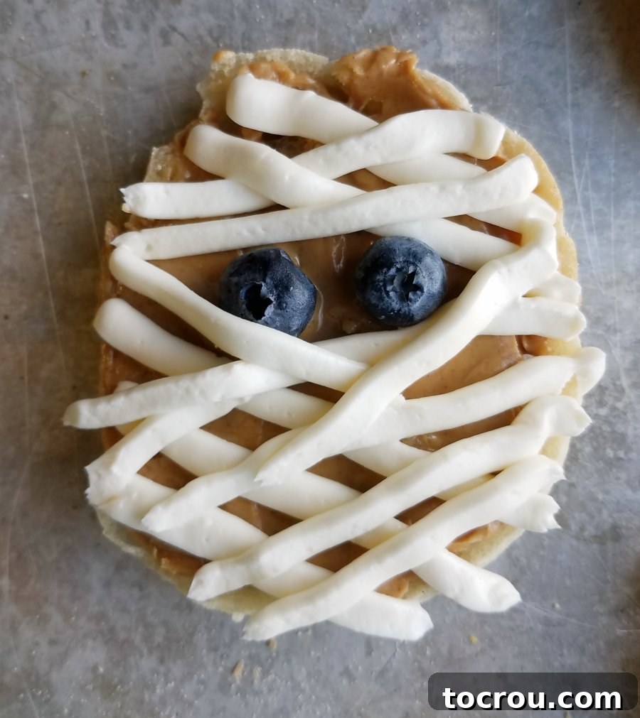 Detailed View of Peanut Butter English Muffin Mummy Close-up of a single English muffin mummy with peanut butter base and detailed cream cheese bandages and blueberry eyes.