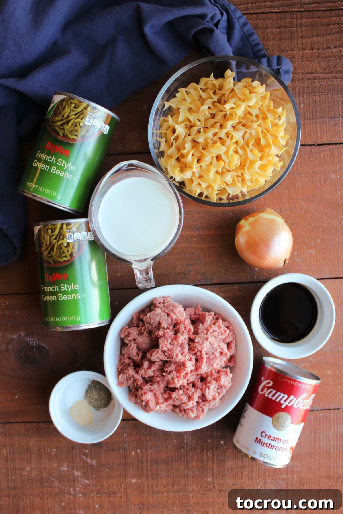 Ingredients including ground beef, an onion, cans of green beans, milk, Worcestershire sauce, garlic, pepper, cream of mushroom soup, and egg noodles ready to be made into a one pot casserole.