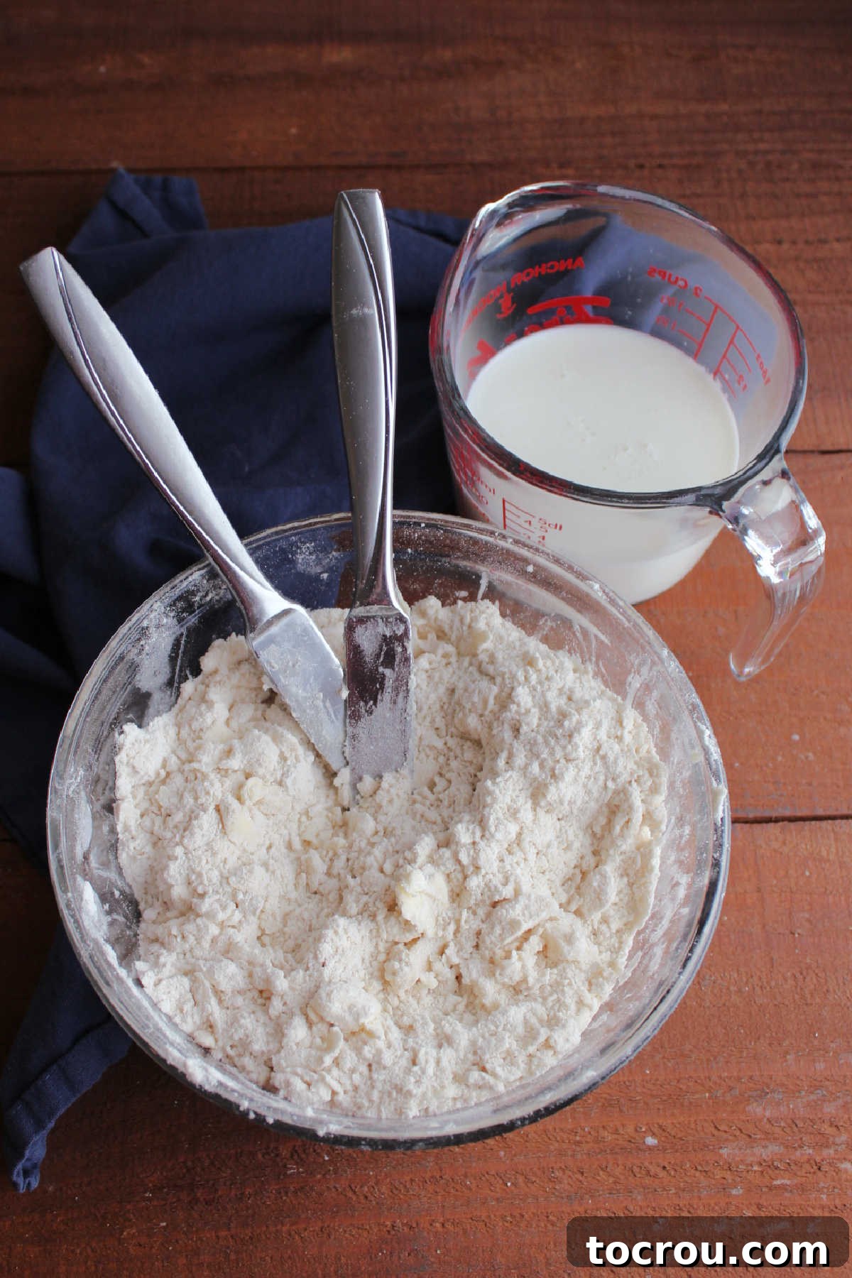 Bowl with butter and lard cut into the cry ingredients to make a crumbly mixture next to a measuring cup with soured milk inside.