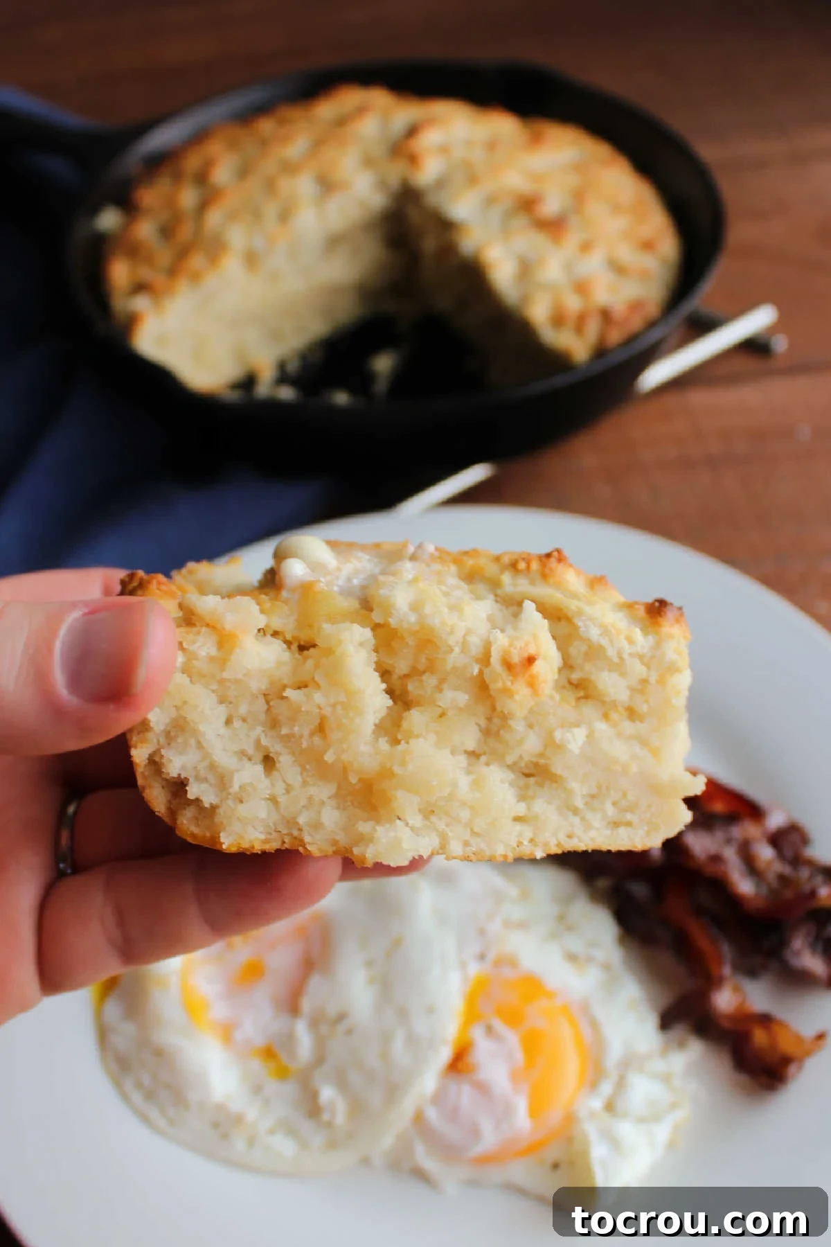 Hand holding piece of pone bread with plate of eggs and bacon in the background.