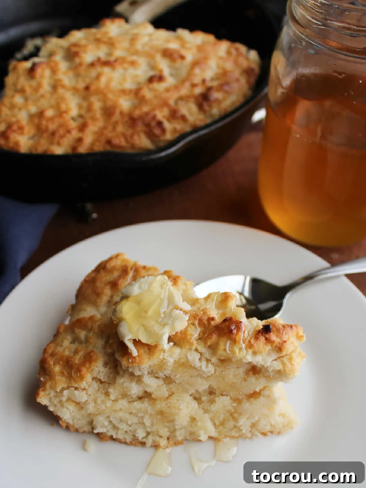 Slice of pone bread topped with butter and honey with cast iron skillet containing more bread and a jar of honey in the background.