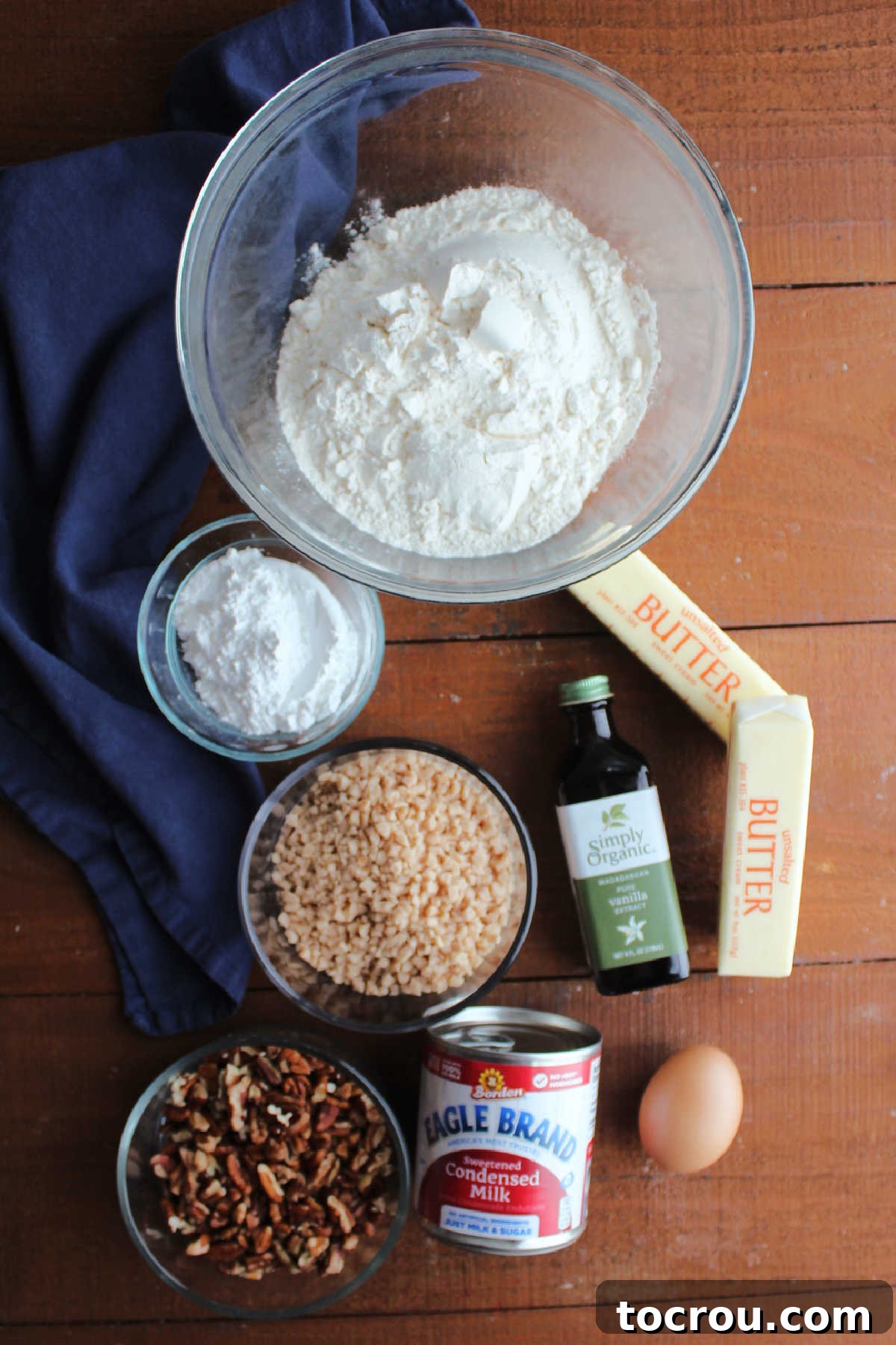 A carefully arranged display of essential ingredients for making pecan pie bars, including all-purpose flour, powdered sugar, softened butter, a can of sweetened condensed milk, a fresh egg, a bottle of vanilla extract, a bowl of chopped pecans, and a small pile of toffee bits.