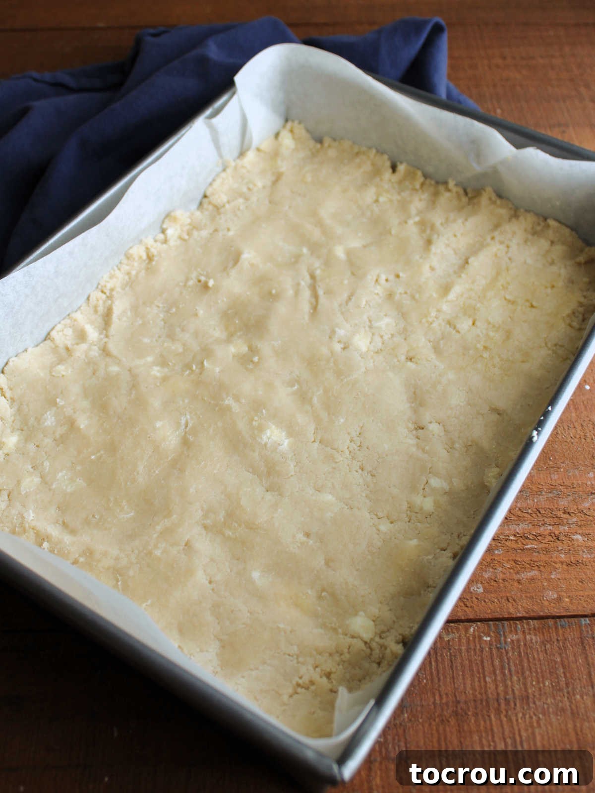 The shortbread crust mixture firmly pressed into an even layer across the bottom of a 9x13-inch baking pan, demonstrating how the loose crumbs come together to form a cohesive, solid base.