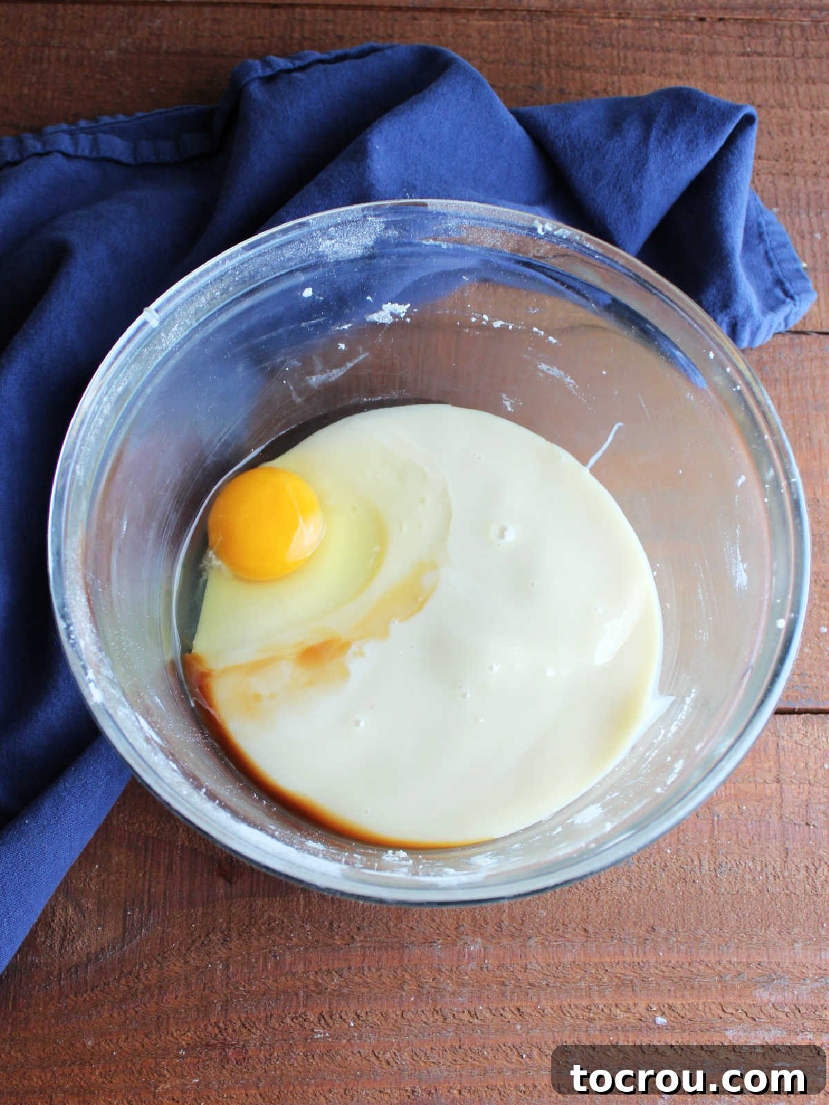 A mixing bowl containing the liquid ingredients for the pecan filling: a can of sweetened condensed milk, a freshly cracked egg, and a clear stream of vanilla extract.