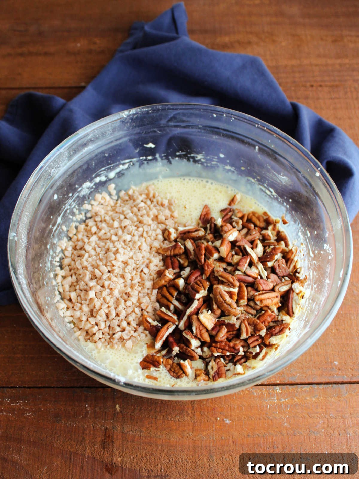 Chopped pecan pieces and small toffee bits being stirred into the smooth, creamy condensed milk and egg mixture, illustrating the creation of the rich and textured pecan pie filling.