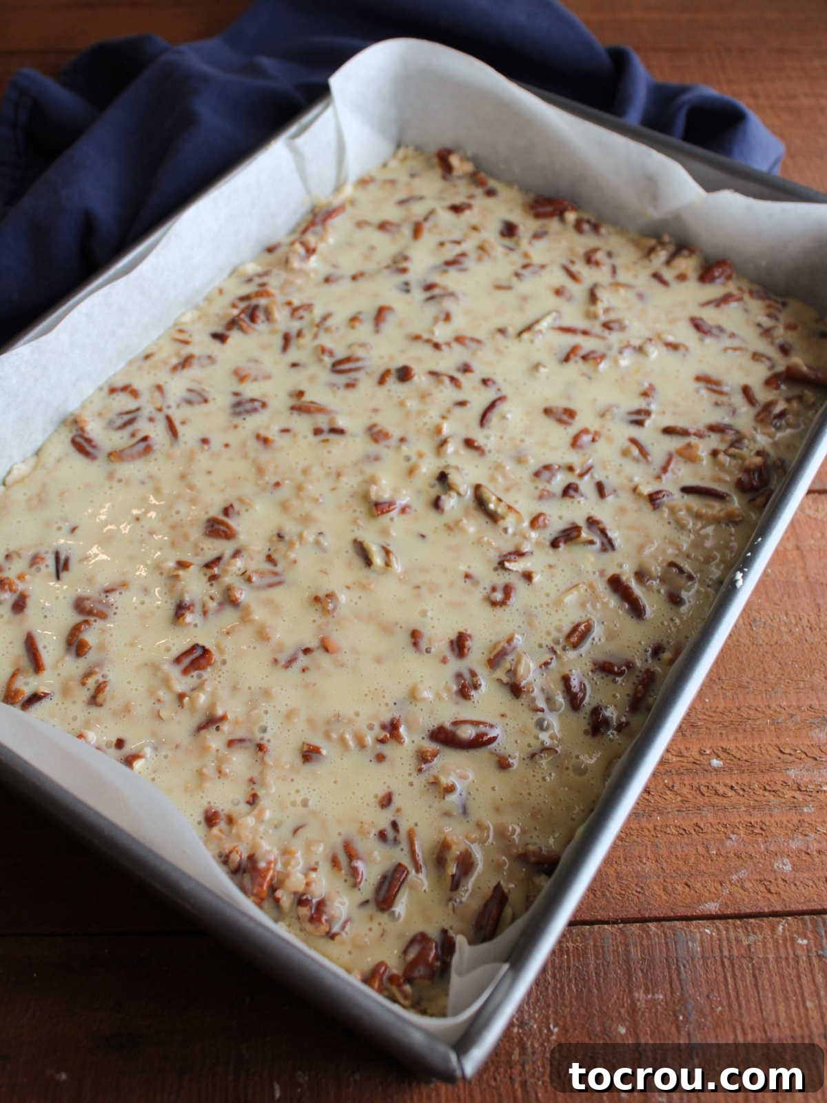 The glistening mixture of sweetened condensed milk and chopped pecans evenly spread over the prepared shortbread crust dough in a baking pan, showing it's ready to be transferred to the oven for baking.
