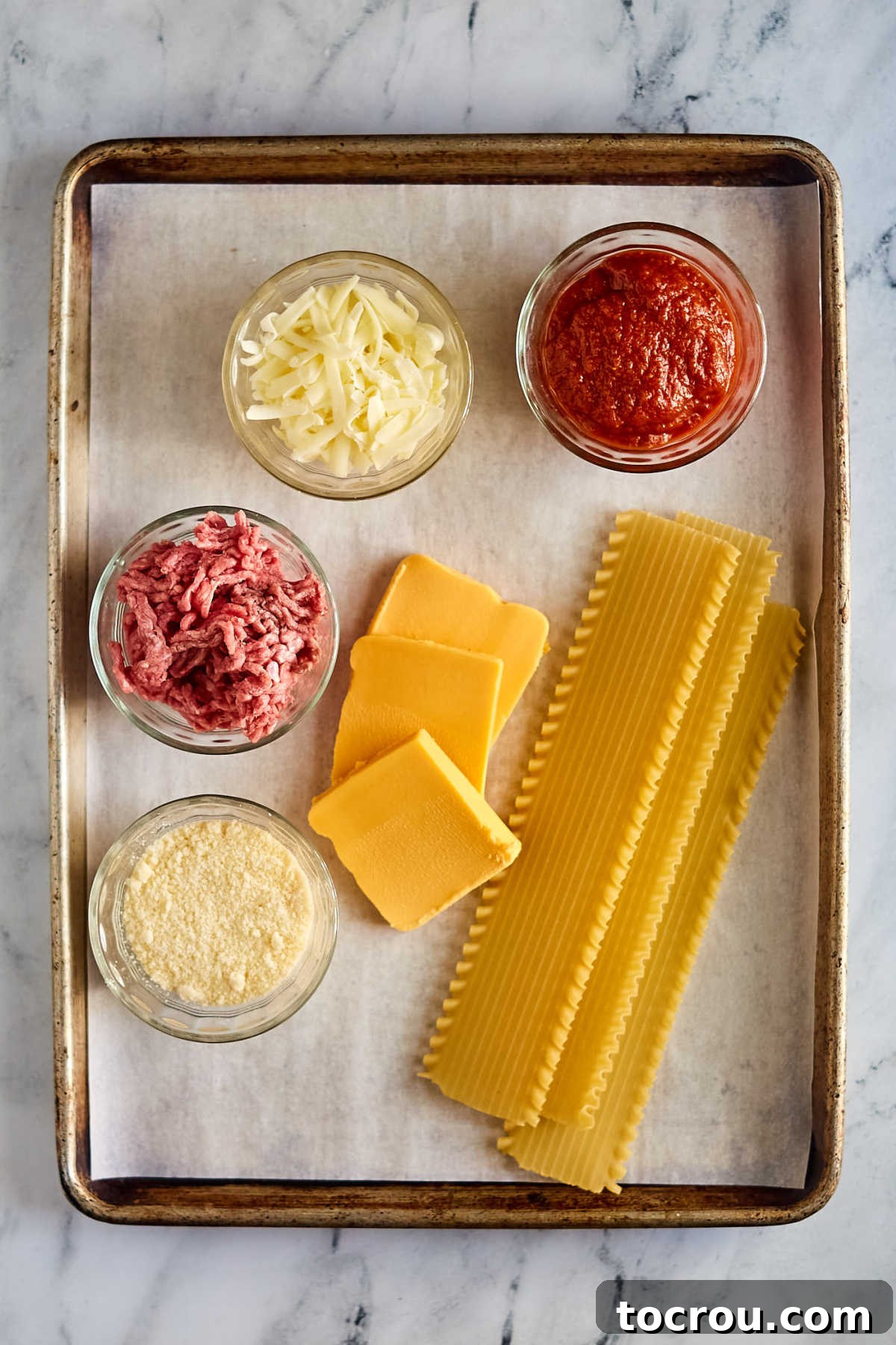 Ingredients for Velveeta Lasagna An overhead shot showcasing the key ingredients for Maw-Maw's Velveeta Lasagna: uncooked lasagna noodles, two jars of rich tomato sauce, a block of Velveeta cheese, shredded mozzarella, lean ground beef, and grated Parmesan cheese, all neatly arranged and ready for preparation.