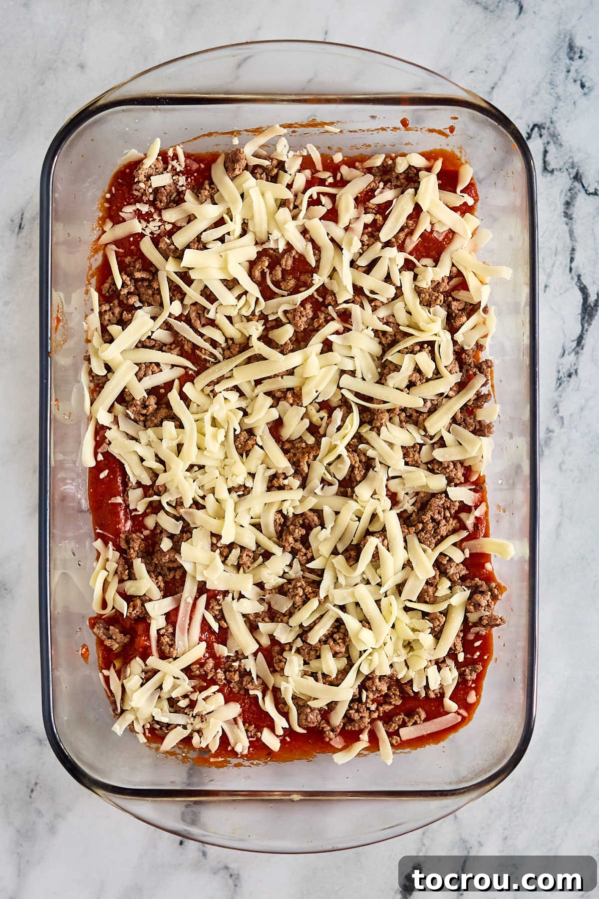 Lasagna Layers: Sauce, Beef, and Mozzarella A glass casserole dish with the initial layers of Maw-Maw's Velveeta lasagna recipe: a base of marinara sauce, a generous scattering of browned ground beef, and a layer of shredded mozzarella cheese, preparing for the next pasta layer.