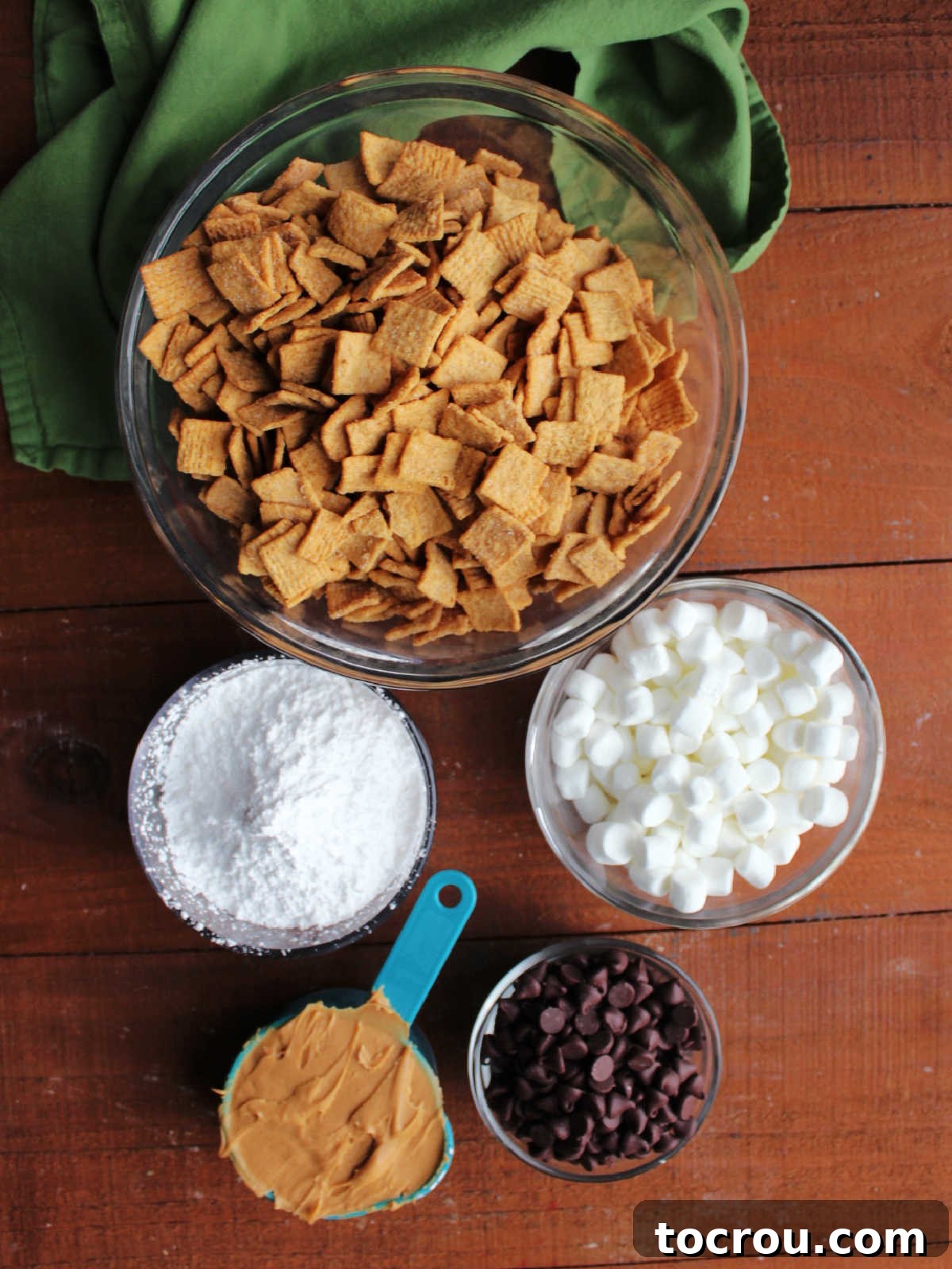 Ingredients including gold grahams cereal, mini marshmallows, chocolate chips, peanut butter, and powdered sugar ready to be made into smores muddy buddies.