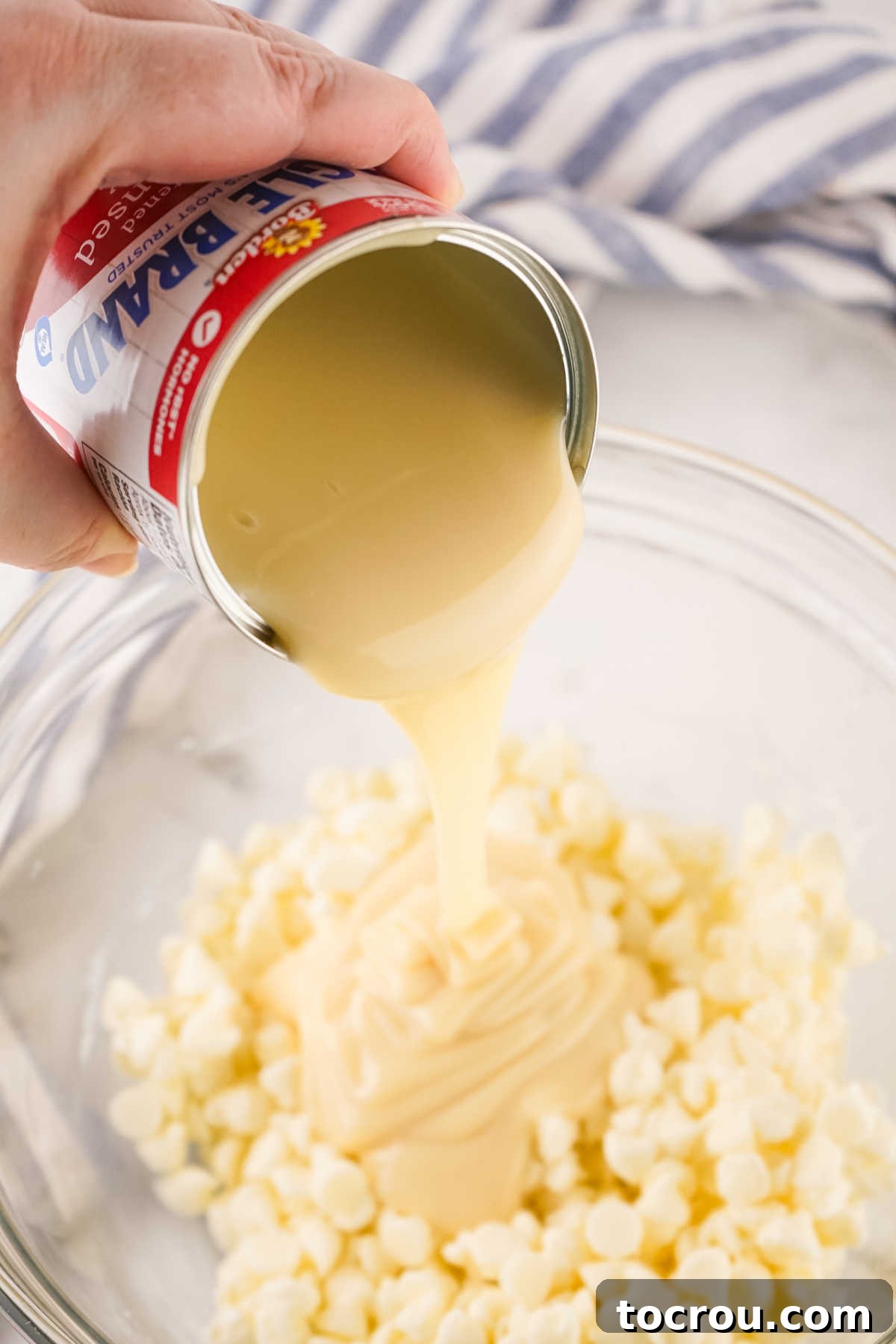 A rich, thick stream of sweetened condensed milk is being poured into a bowl brimming with white chocolate chips, the foundation of this delectable fudge.