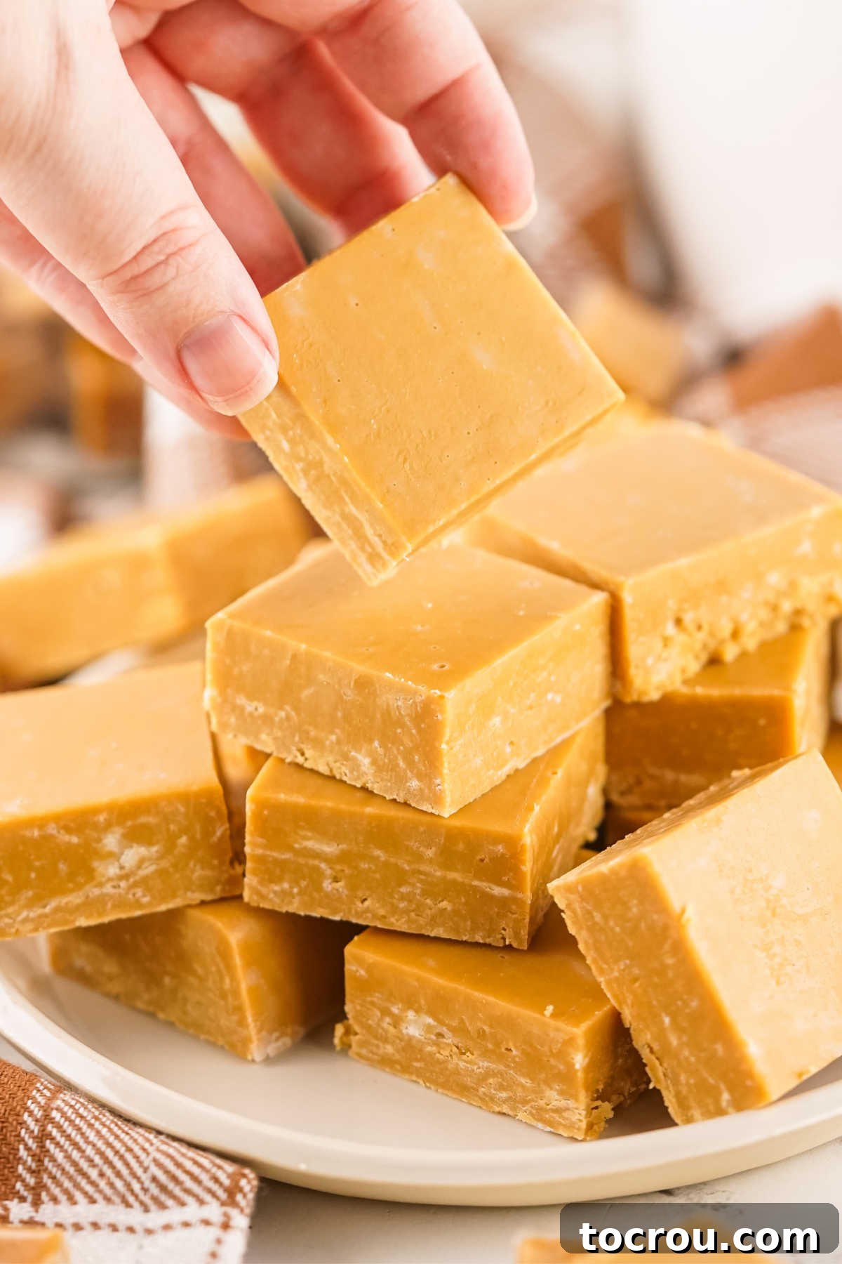 Homemade Caramel Fudge Hand-Held Hand holding square of caramel fudge with plate of more fudge in the background.